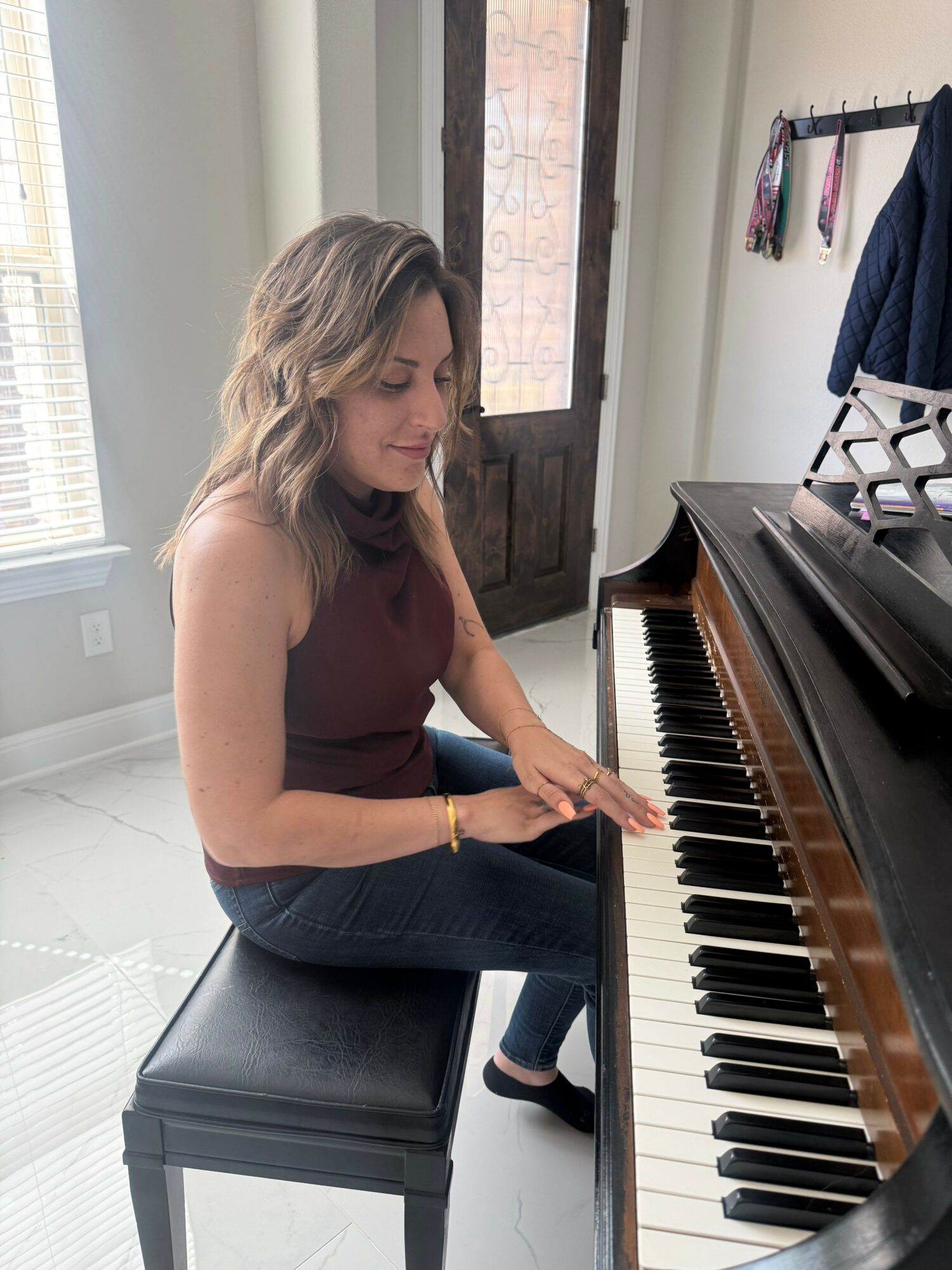 Woman playing piano in a bright room with large windows, sitting on a bench, with a photo on the wall.
