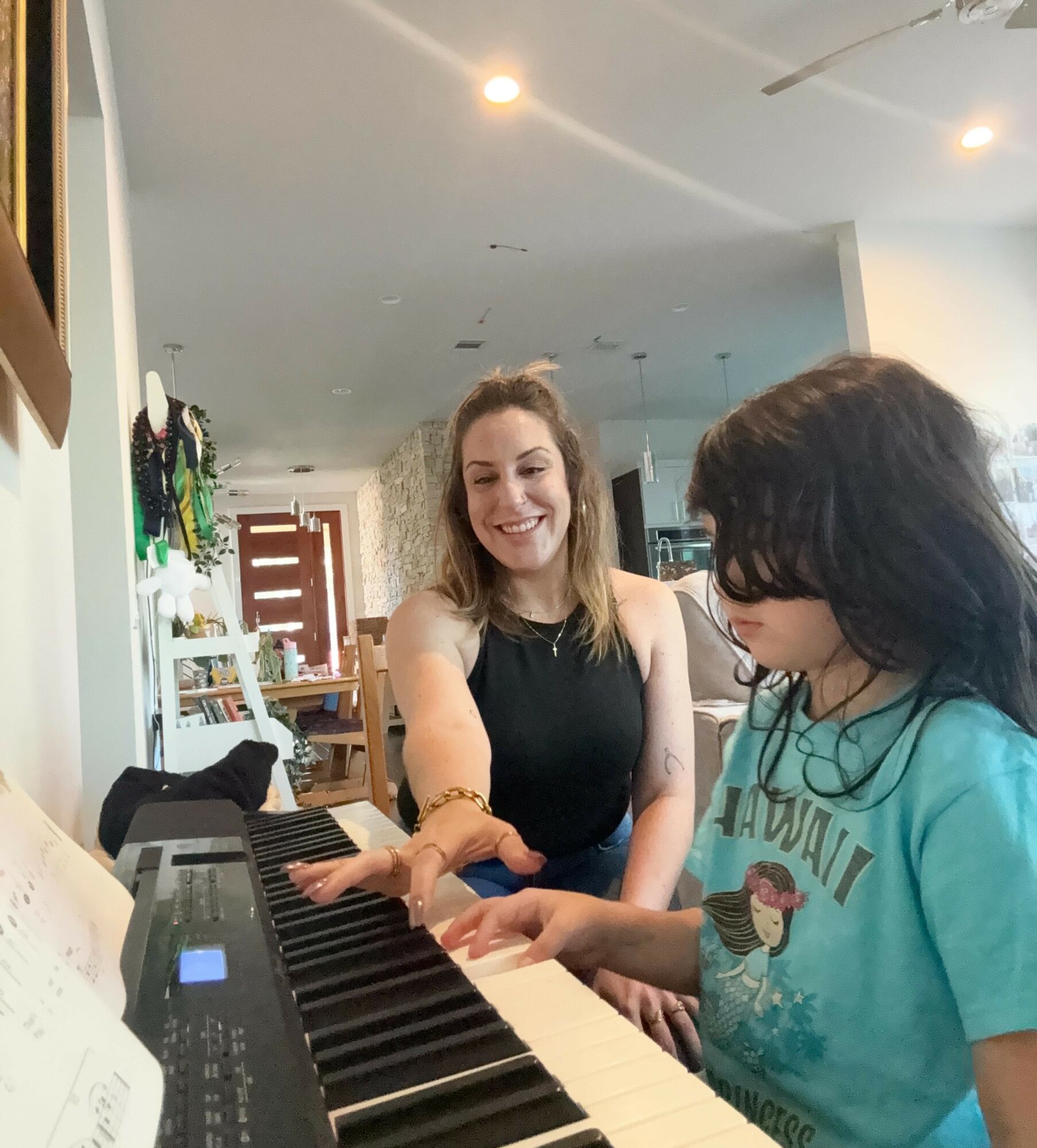 Two girls, one smiling and the other playing a keyboard, in a room with bright lighting and furniture.
