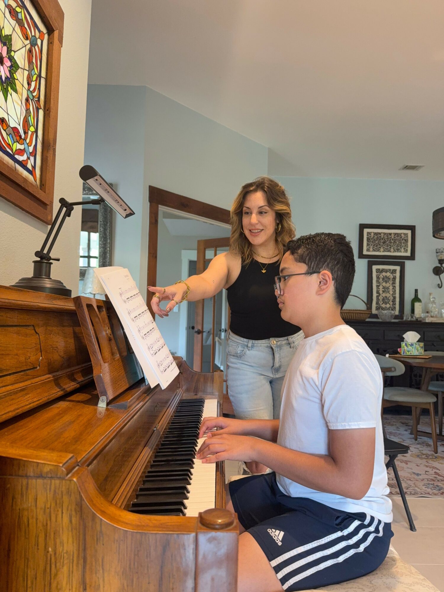 Two people, a woman and a young man, sit at a piano in a room with framed pictures on the wall, engaging with sheet music.