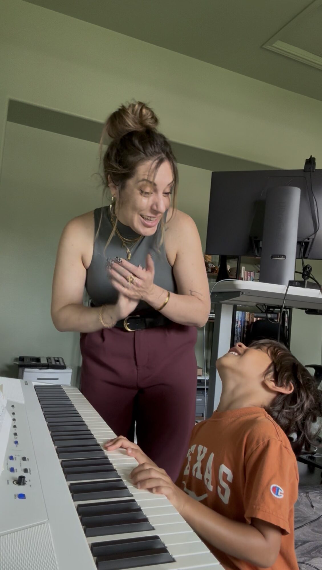 A woman and a girl are smiling and laughing together in a music studio, with a keyboard and computer visible.