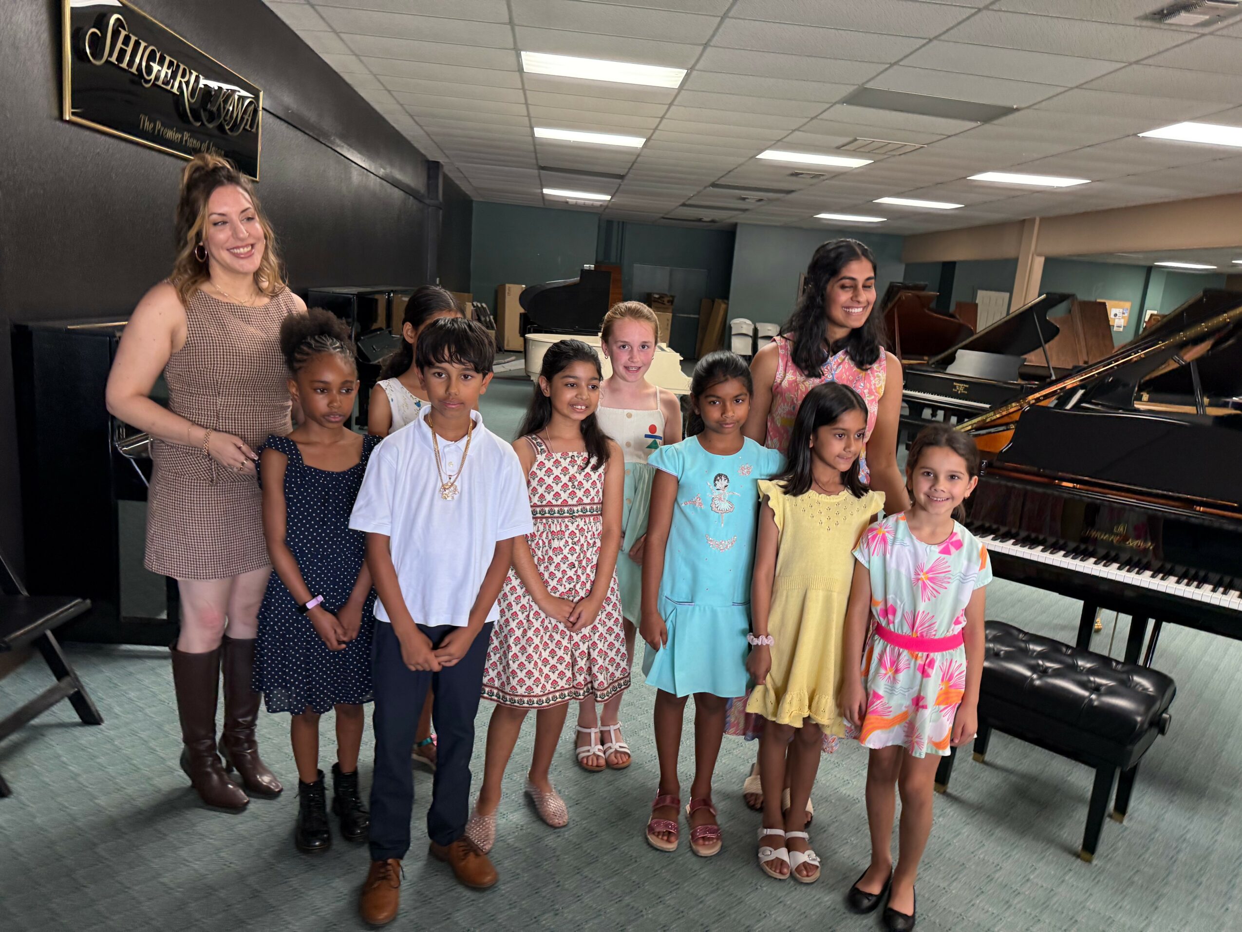 Group of children and an adult woman standing in a music room with pianos, smiling, dressed in colorful clothes.
