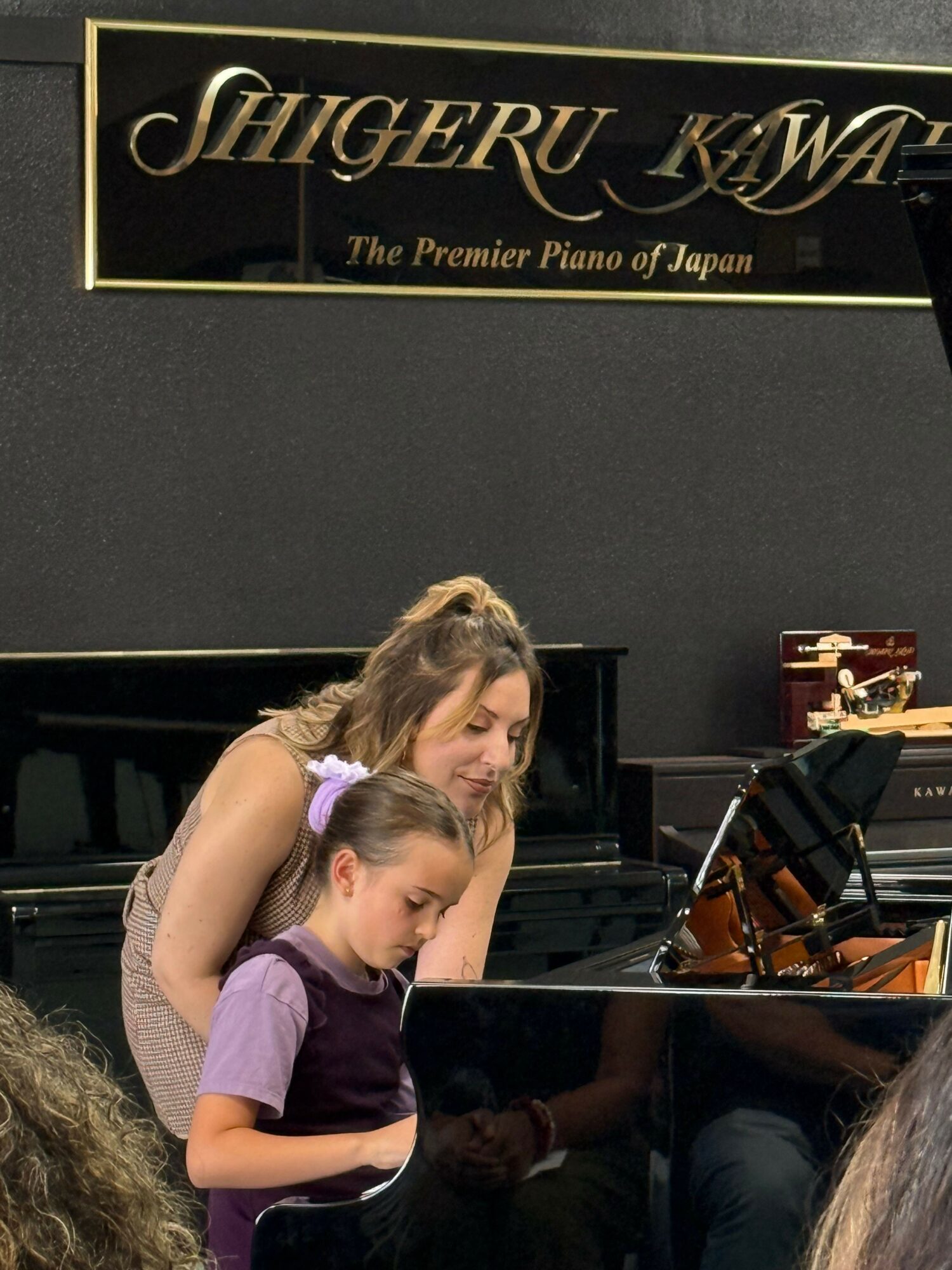 Two young girls sit at a piano, one adjusting the other's hair, in a music room with a sign reading 'Shepherd U.'