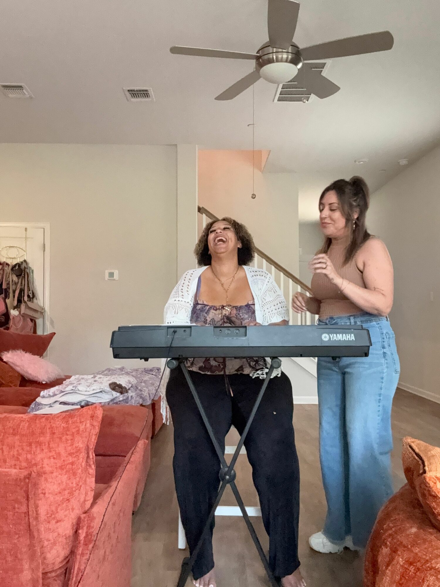 Two women stand in a room, one playing a keyboard, the other watching, with a ceiling fan overhead.