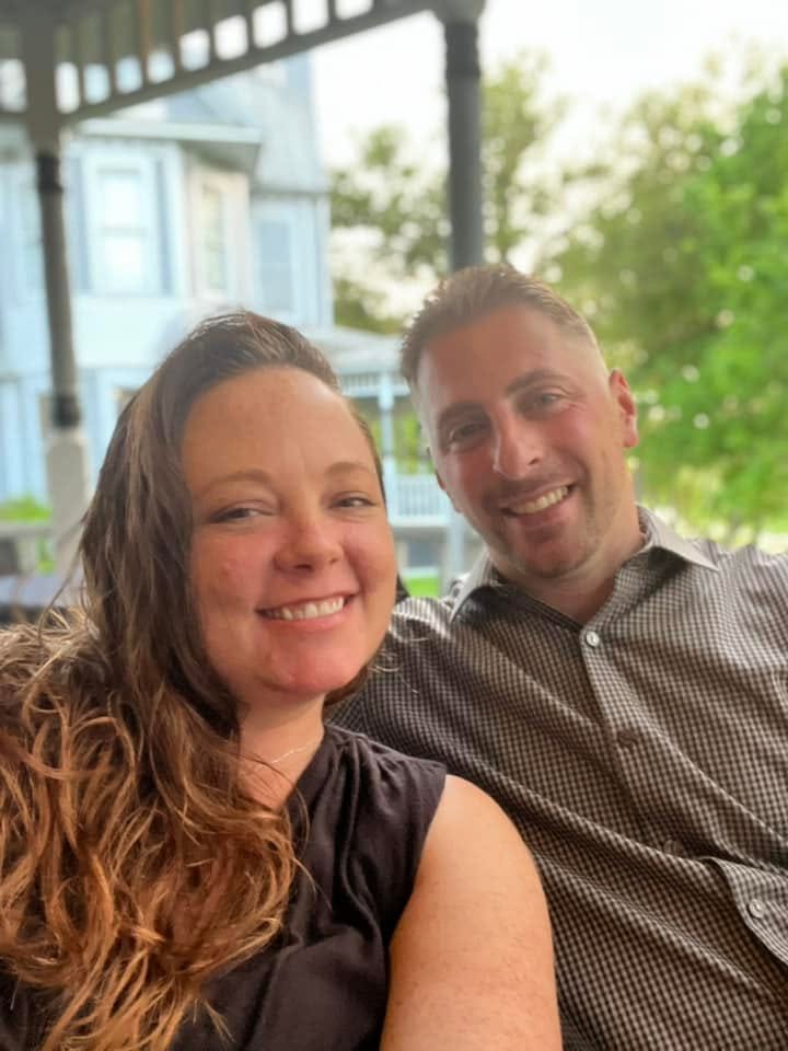 Two people smiling outdoors with trees and a building in the background.