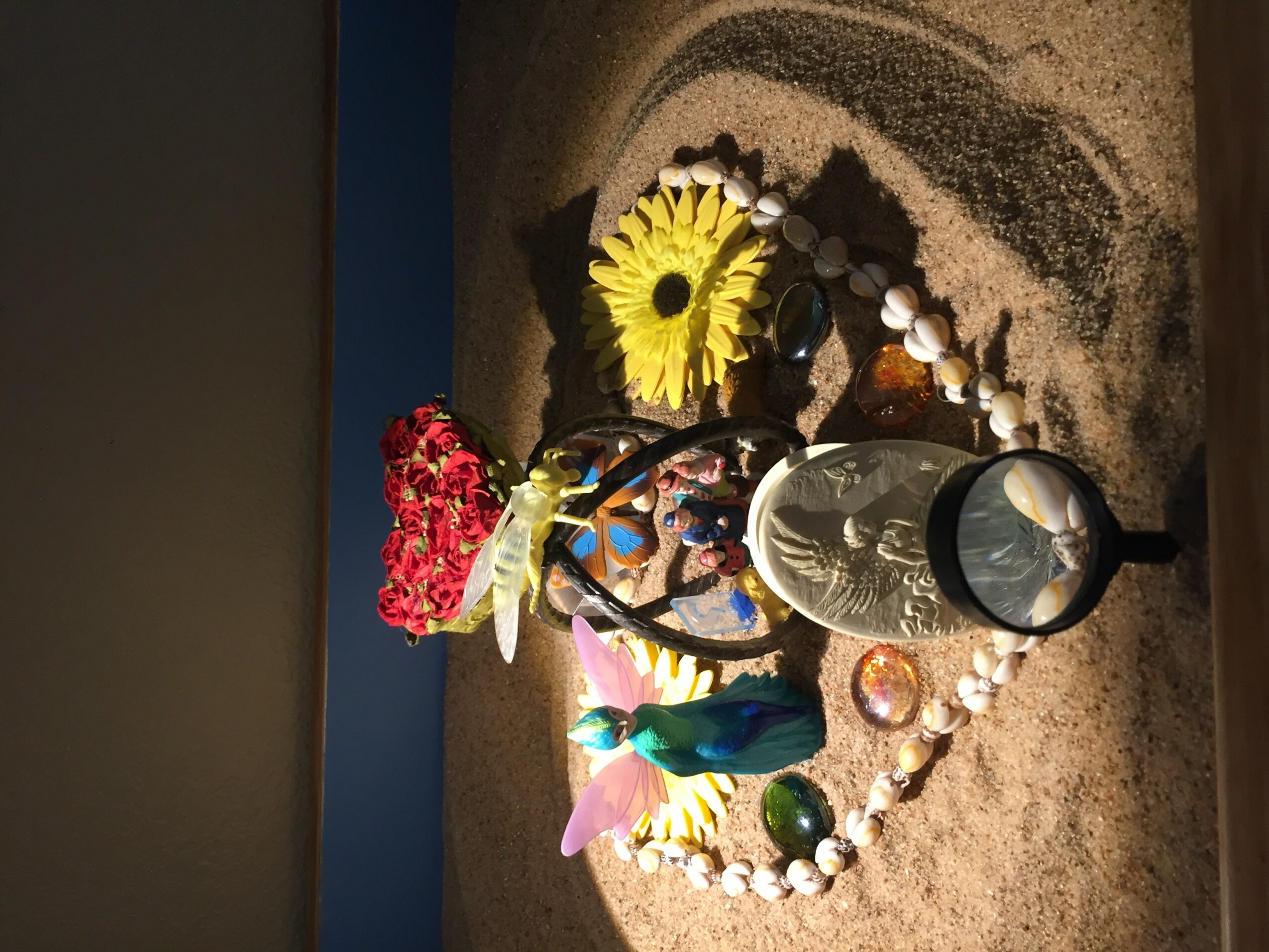 Decorative arrangement with flowers, a glass, and colorful objects on a sandy surface.