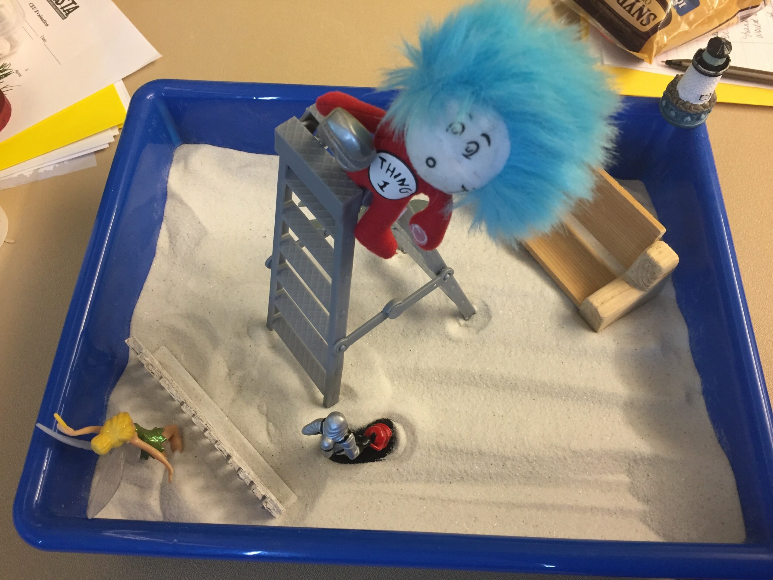 Toy scene with a blue tray filled with white sand, a plush doll with blue hair, a small figure, and various objects.