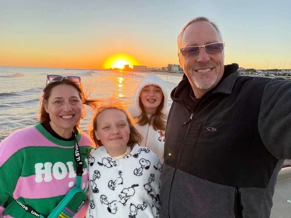 Four people, two children and two adults, smiling at sunset by the water, with the sun near the horizon.