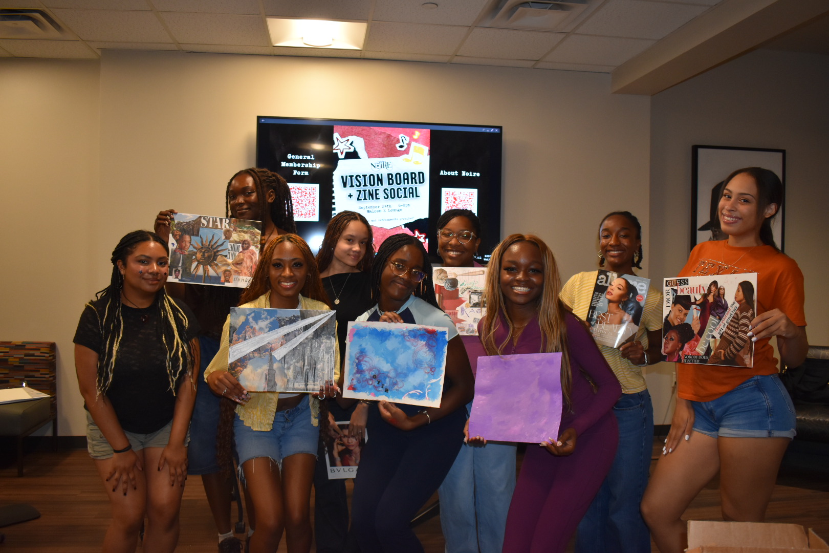 Group of eight young women holding artwork in a room with a screen displaying a presentation slide, smiling.