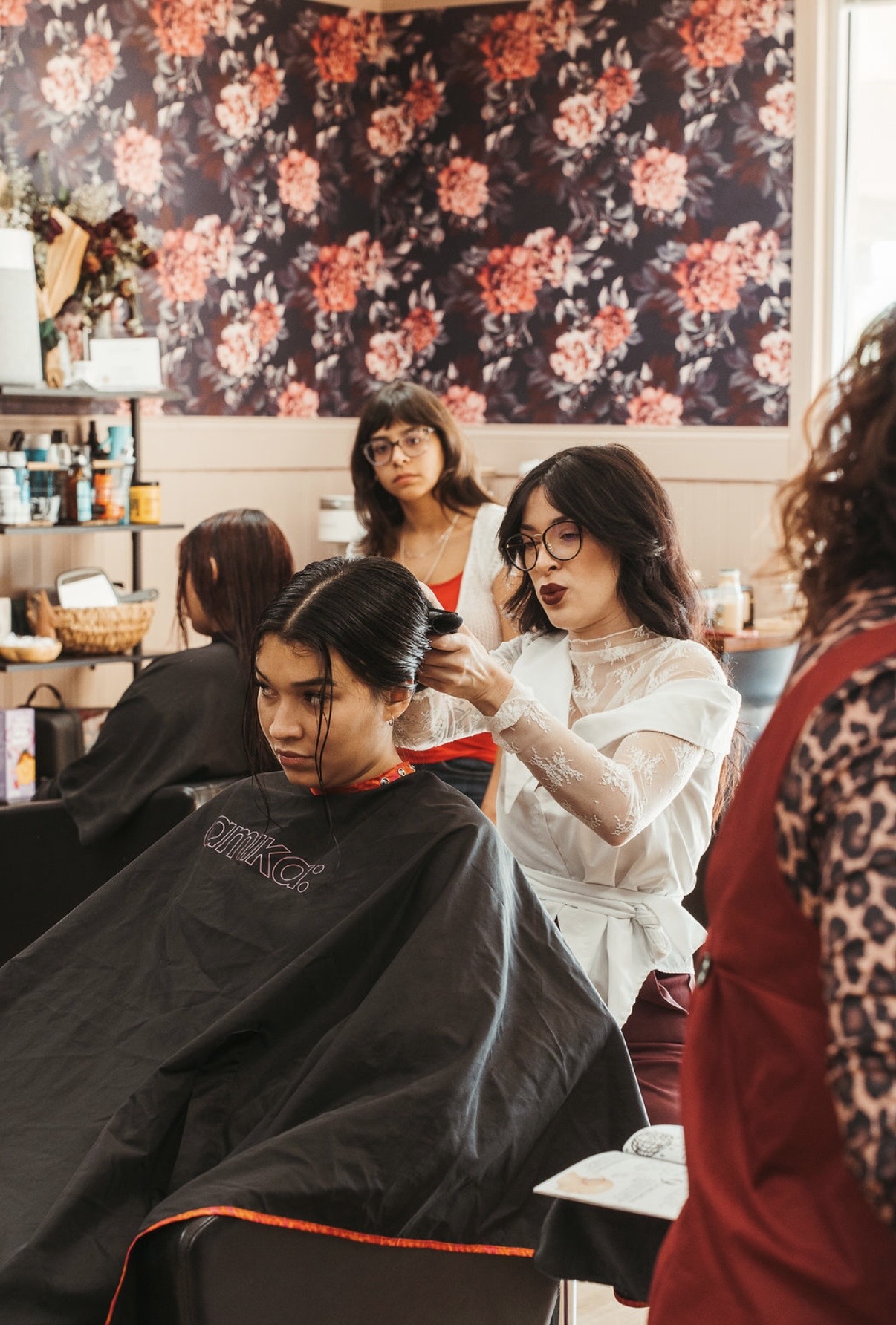 A woman getting her hair styled in a salon with floral wallpaper background, other clients and stylist present.