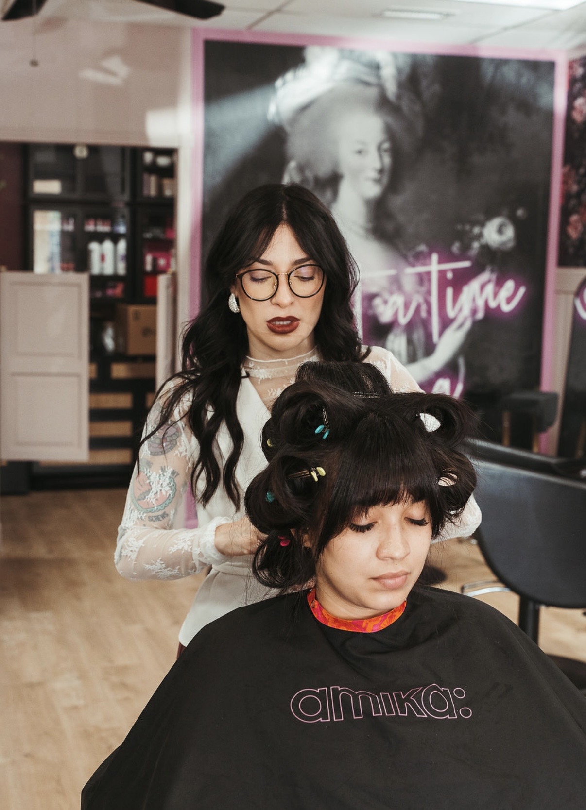 A woman with dark hair and glasses gets her hair styled with rollers in a salon, with a poster in the background.