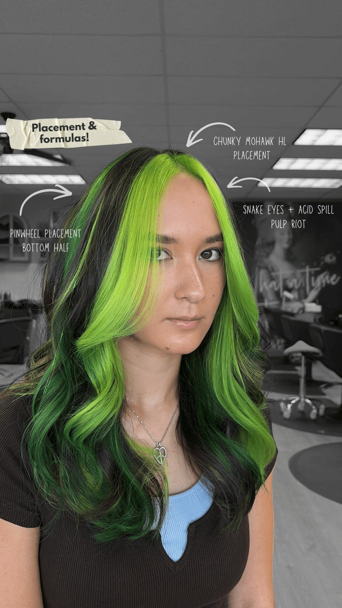Woman with long, wavy hair dyed bright green and black, standing in a classroom or office setting.