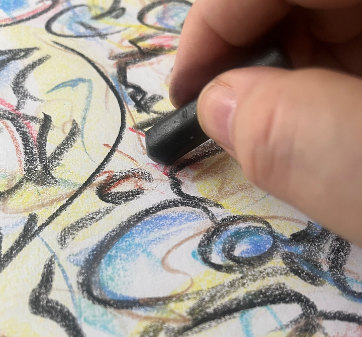 Close-up of a hand holding a black crayon drawing colorful abstract shapes on paper.