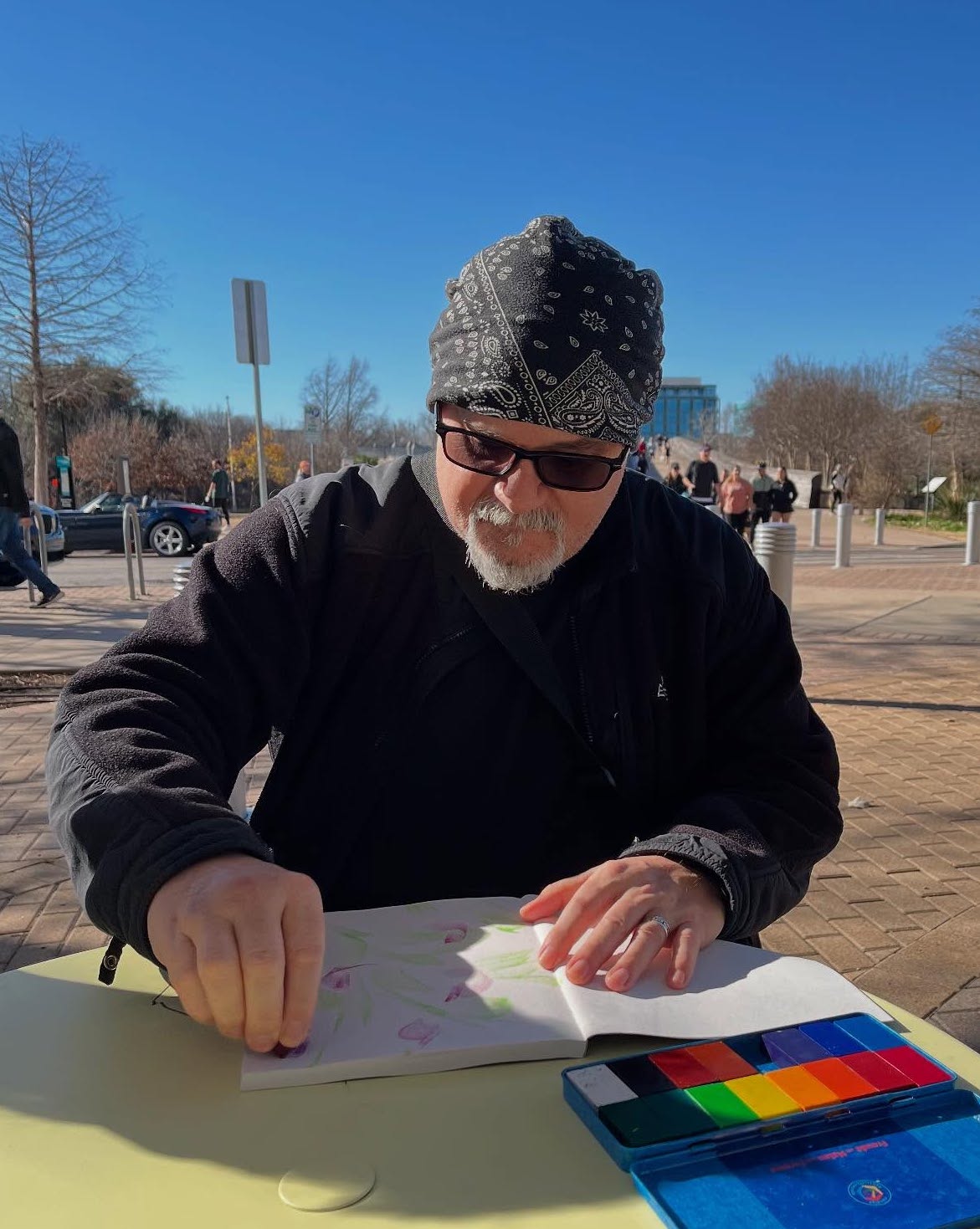 Man wearing sunglasses, a patterned headscarf, and a black jacket, sitting outdoors at a table with a colorful palette, reading a book.