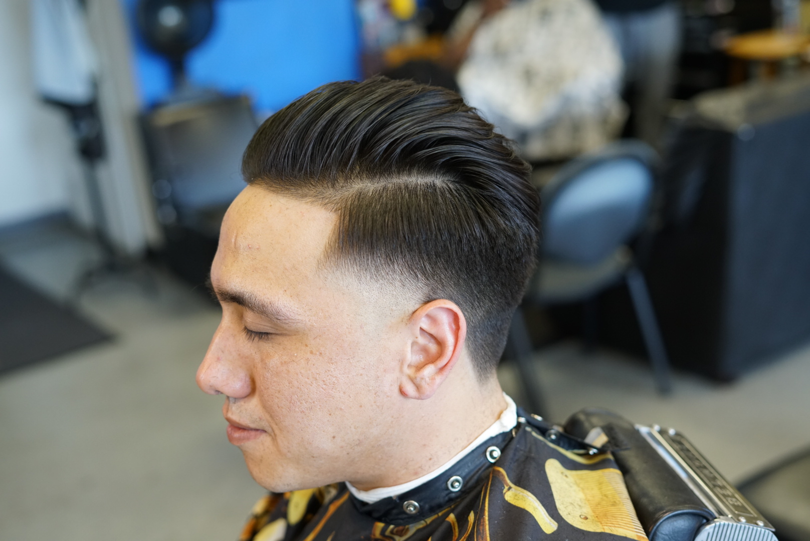 Young man with a styled haircut, side profile, in a barbershop, wearing a cape, background with chairs and people.