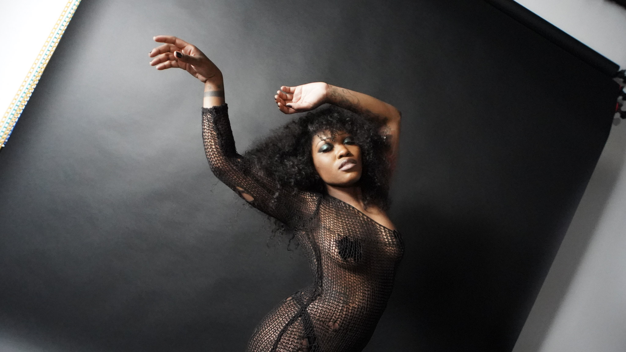 Woman with curly hair in a black fishnet outfit poses against a dark background.