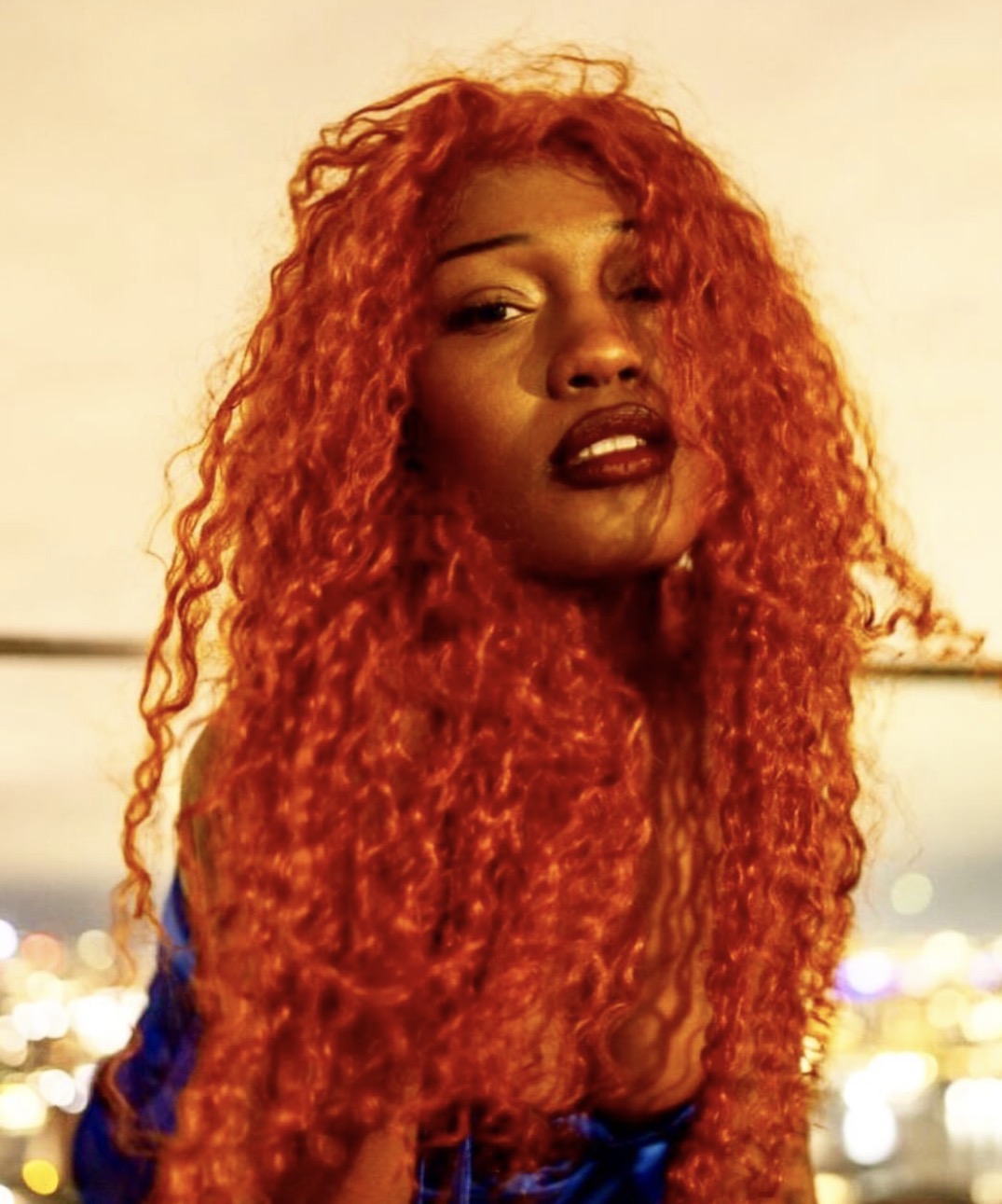 Woman with long, curly, bright red hair and dark lipstick, looking at the camera, background with city lights.