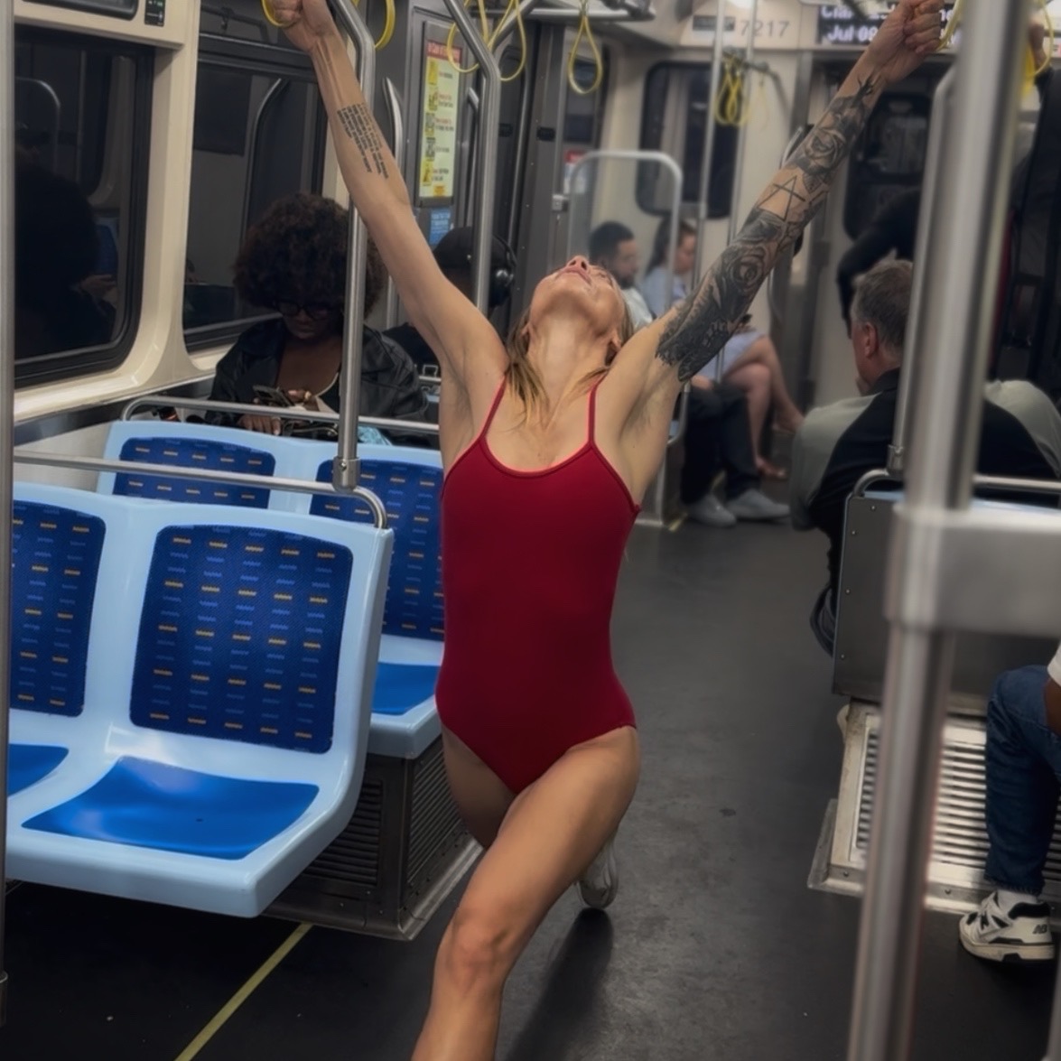 Person in a red swimsuit with tattoos, arms raised, standing inside a subway train, with other passengers seated around.