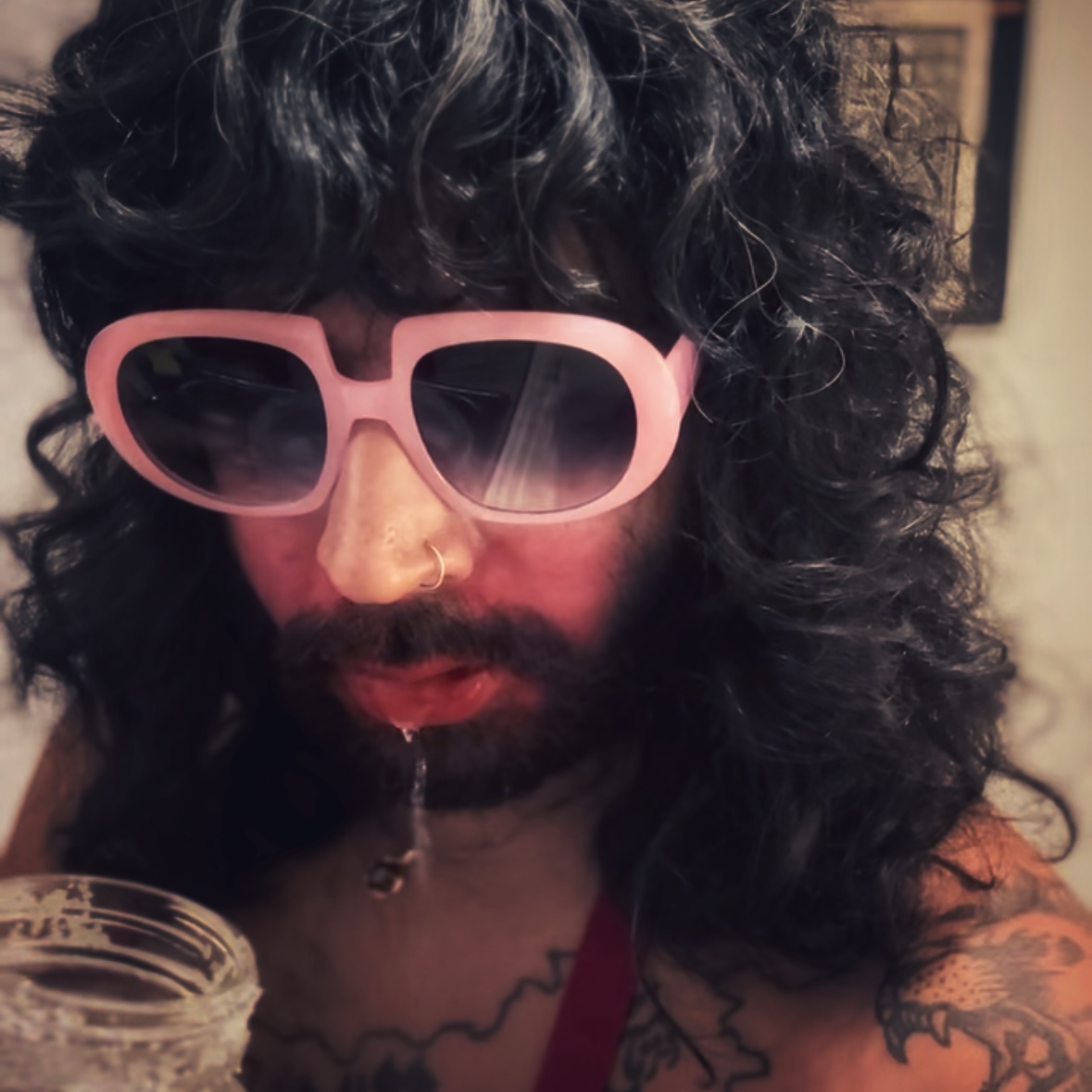 Person with curly hair wearing large pink sunglasses, with tattoos visible on chest and shoulder, holding a glass jar.