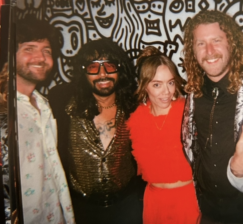 Four people smiling in front of a black and white patterned wall, dressed in colorful and retro clothing.