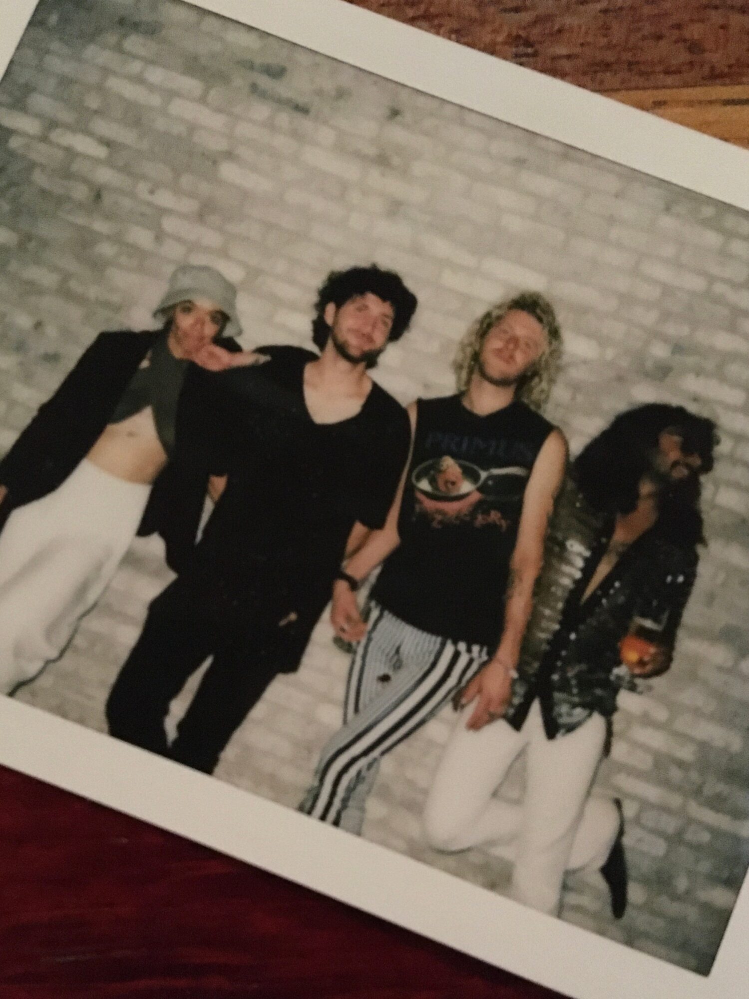 Four people standing against a brick wall, posing for a photo, with casual clothing and accessories.