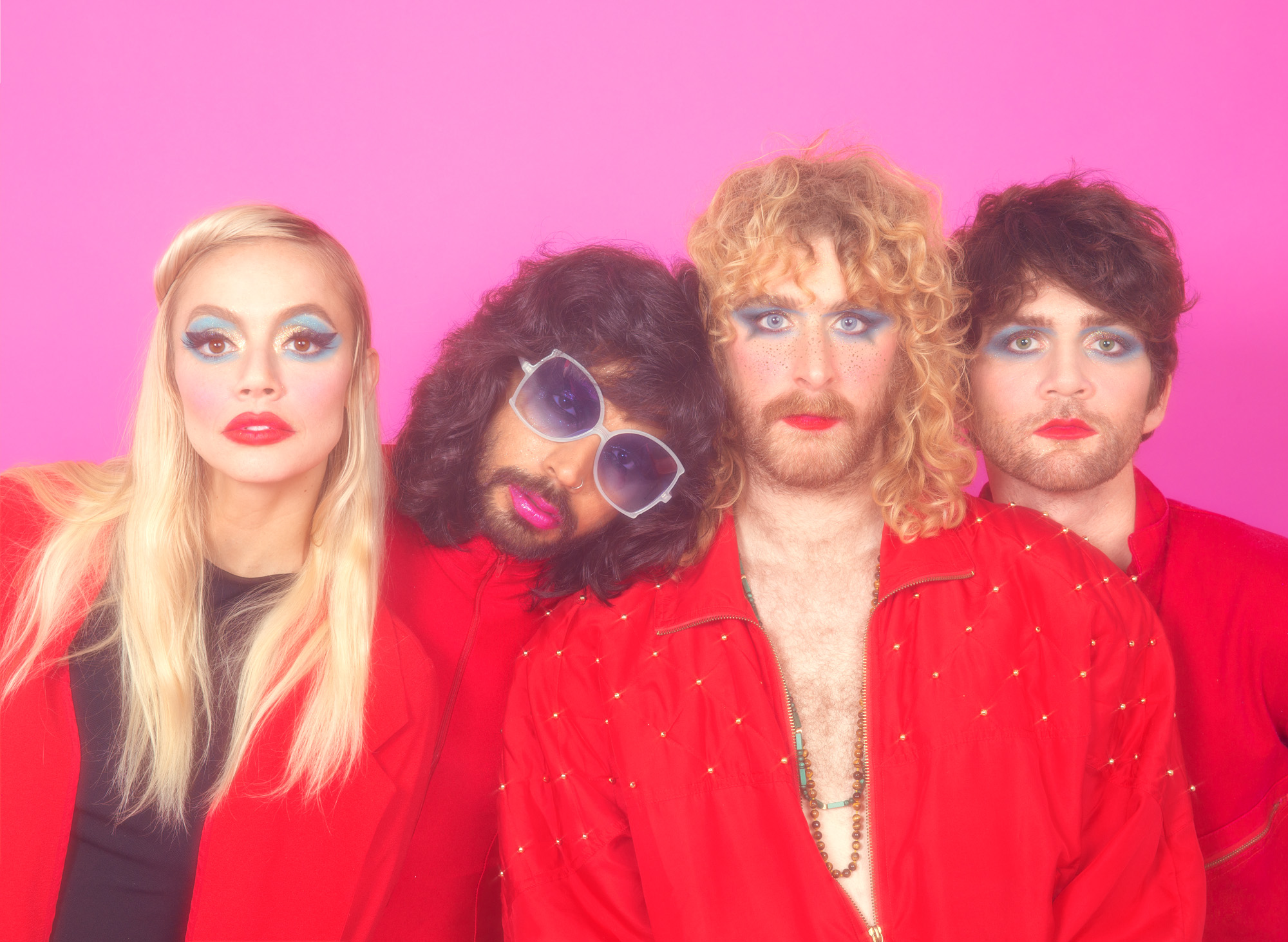 Four people with colorful makeup and curly hair pose against a pink background, wearing red and black clothing.