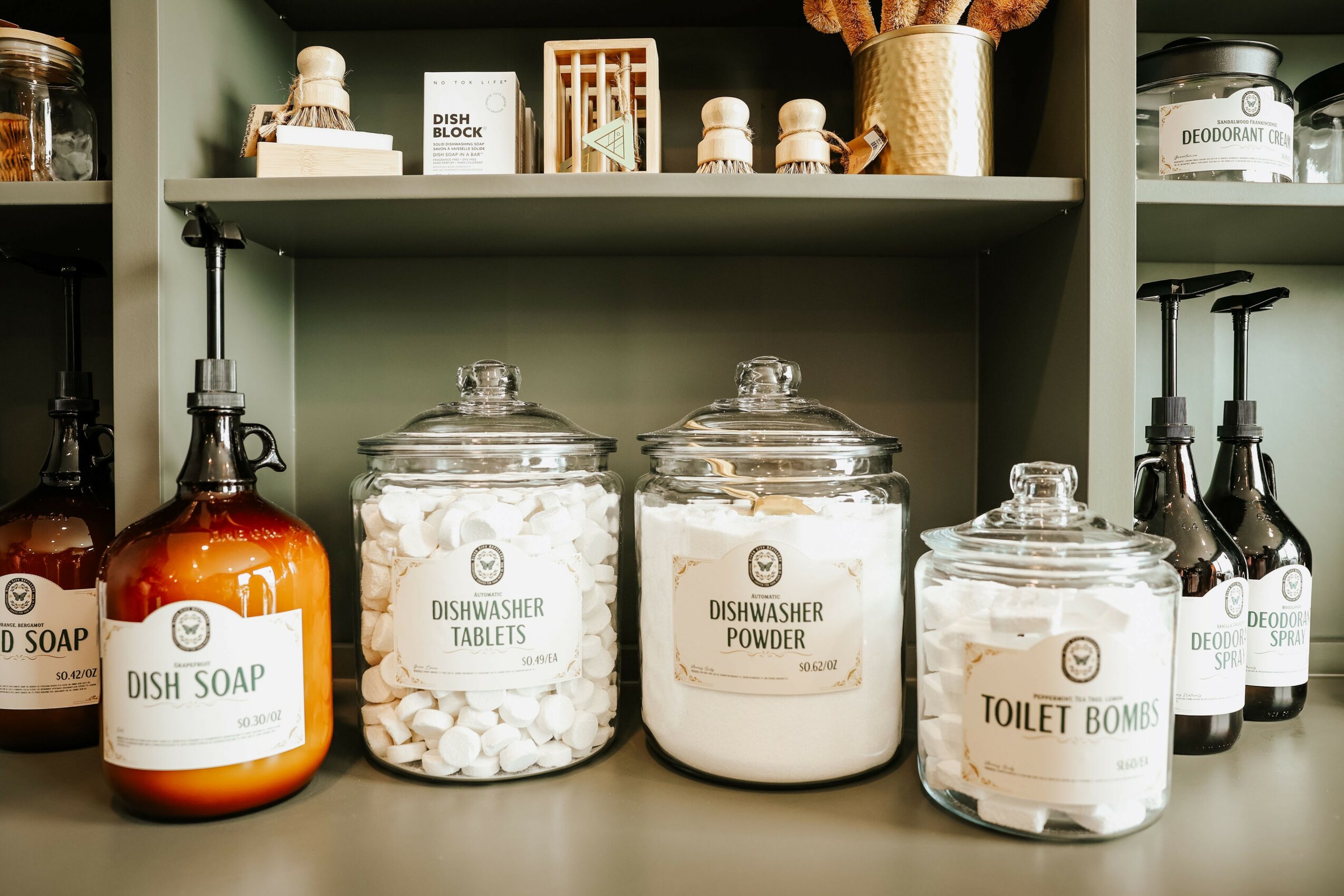Shelves with jars labeled dish soap, dishwasher tablets, dishwasher powder, and toilet bombs, along with small bottles and containers.
