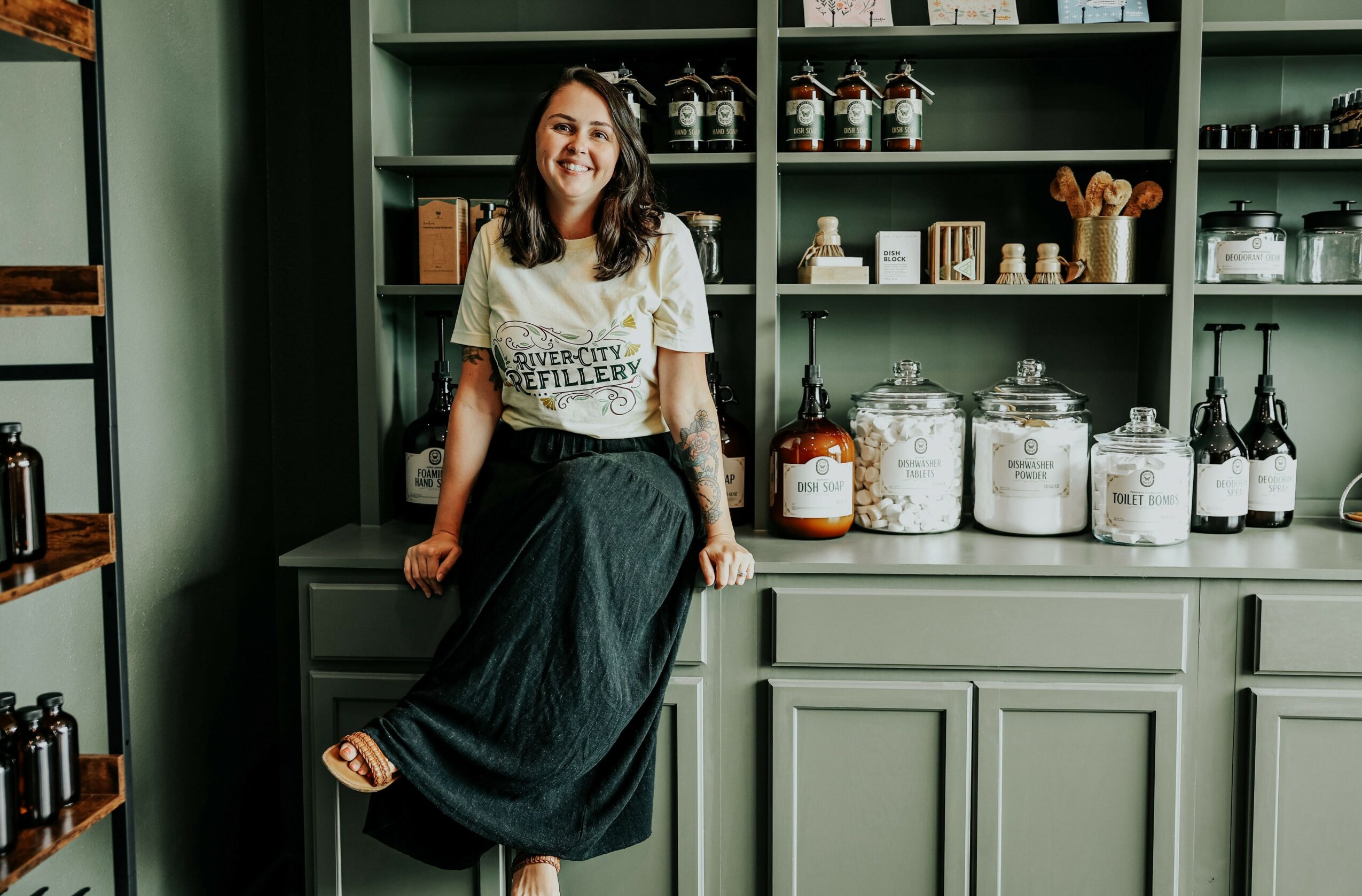 Woman with long hair smiling, sitting on a kitchen counter, surrounded by green cabinets and jars.