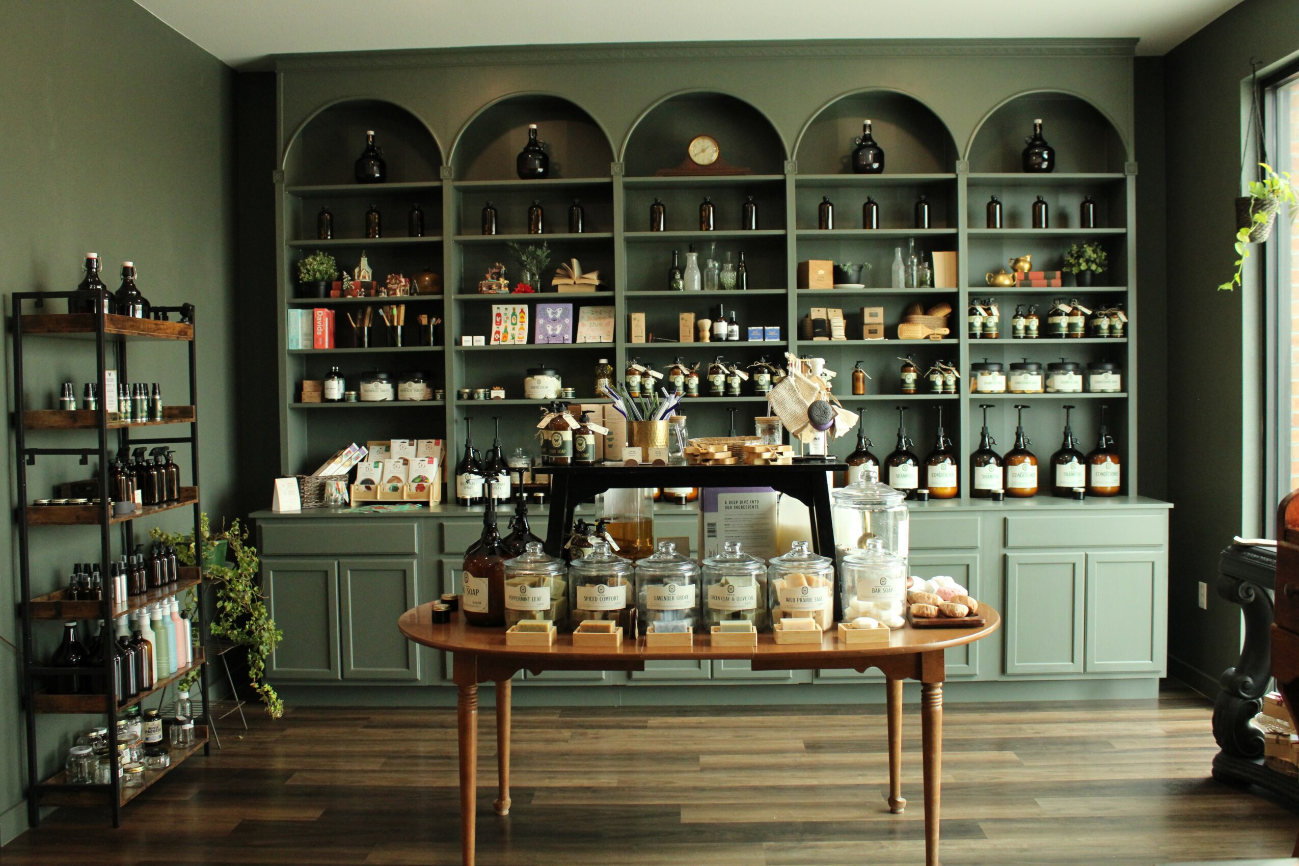 Green display cabinet with shelves filled with jars, bottles, and decorative items, and a wooden table with glass jars in front.