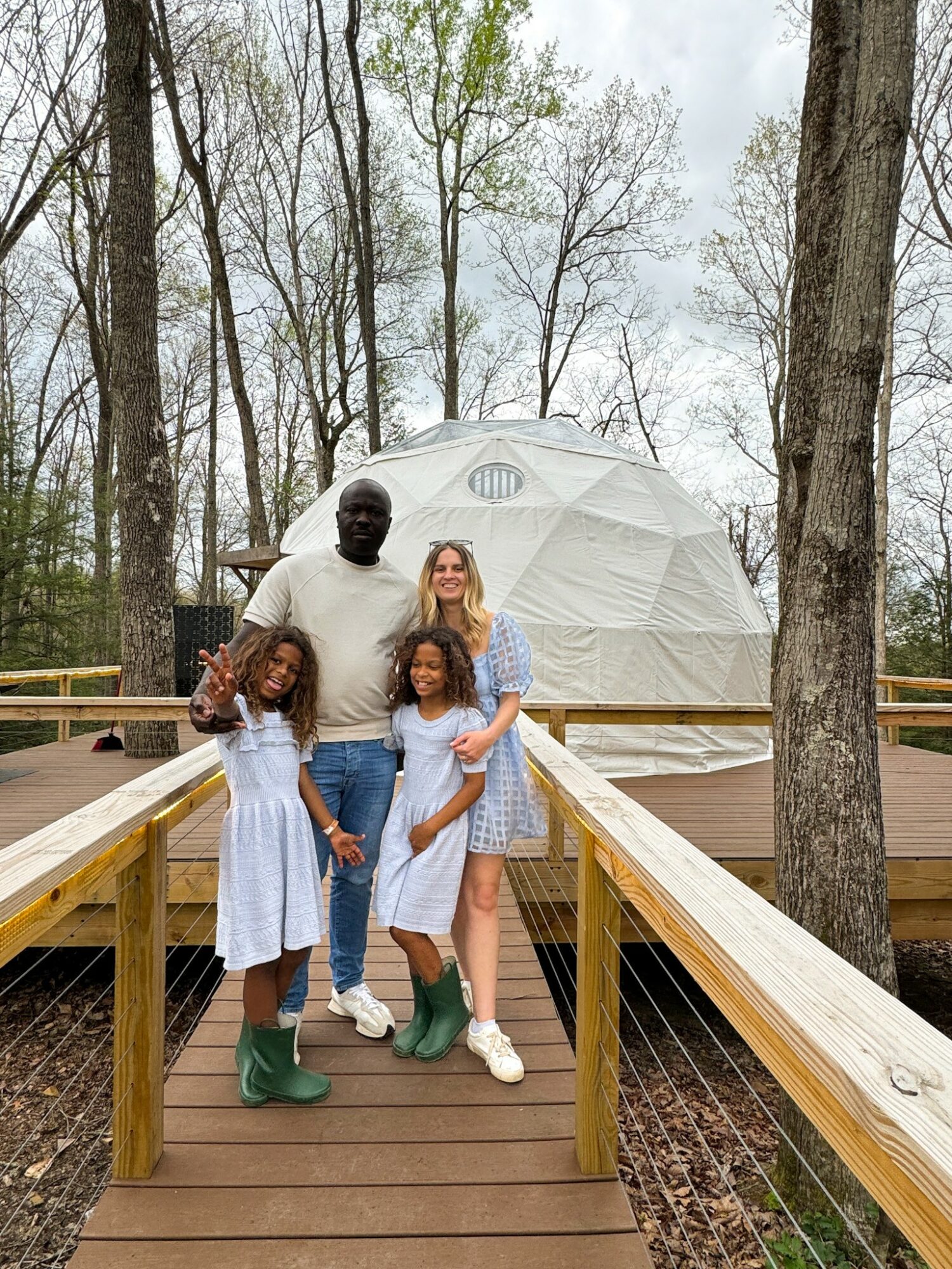 A family of four stands on a wooden bridge in a forest, with a geodesic dome in the background, smiling.