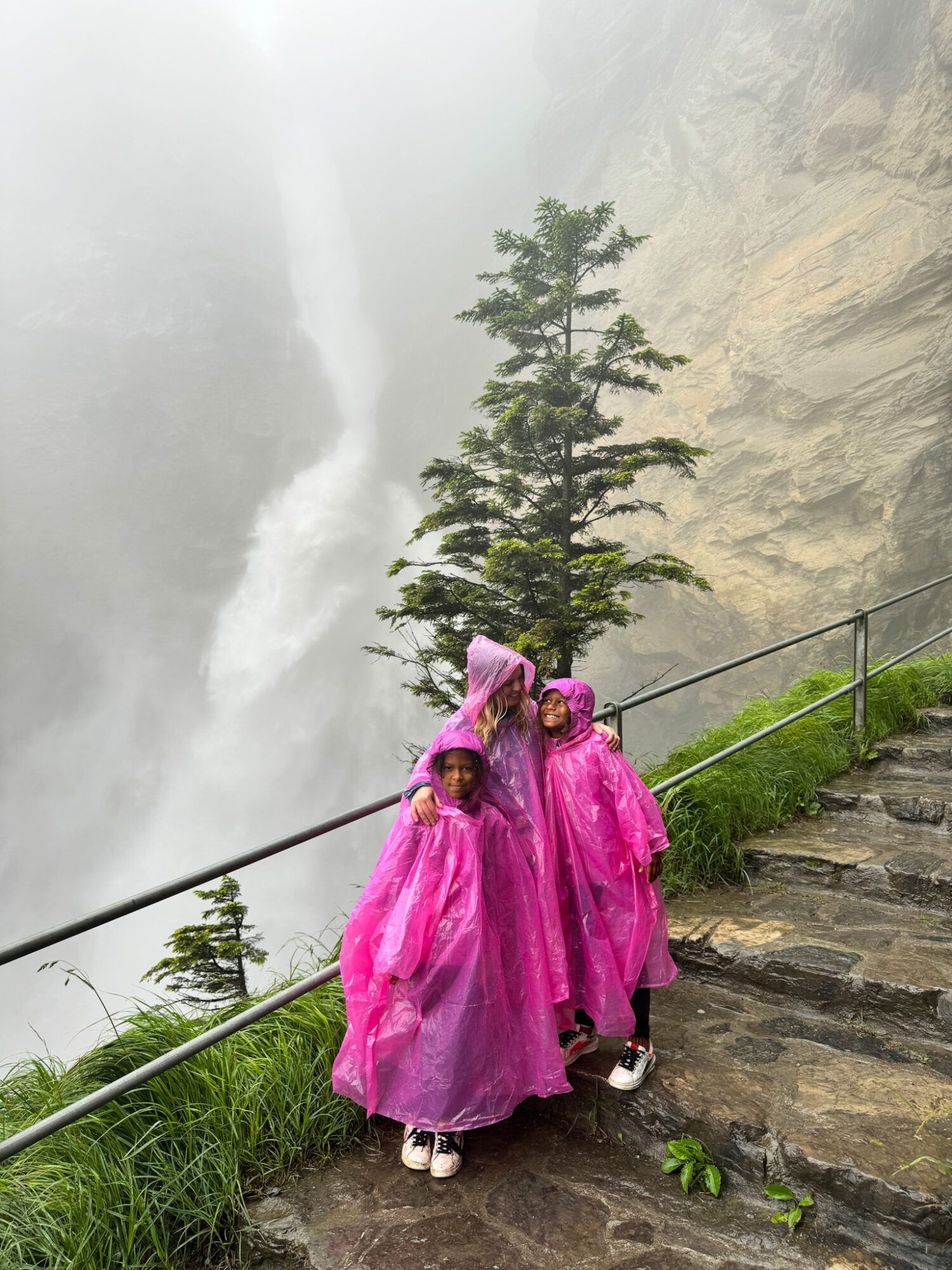 Three people wearing pink rain ponchos standing on a stone path near a waterfall with mist and greenery.