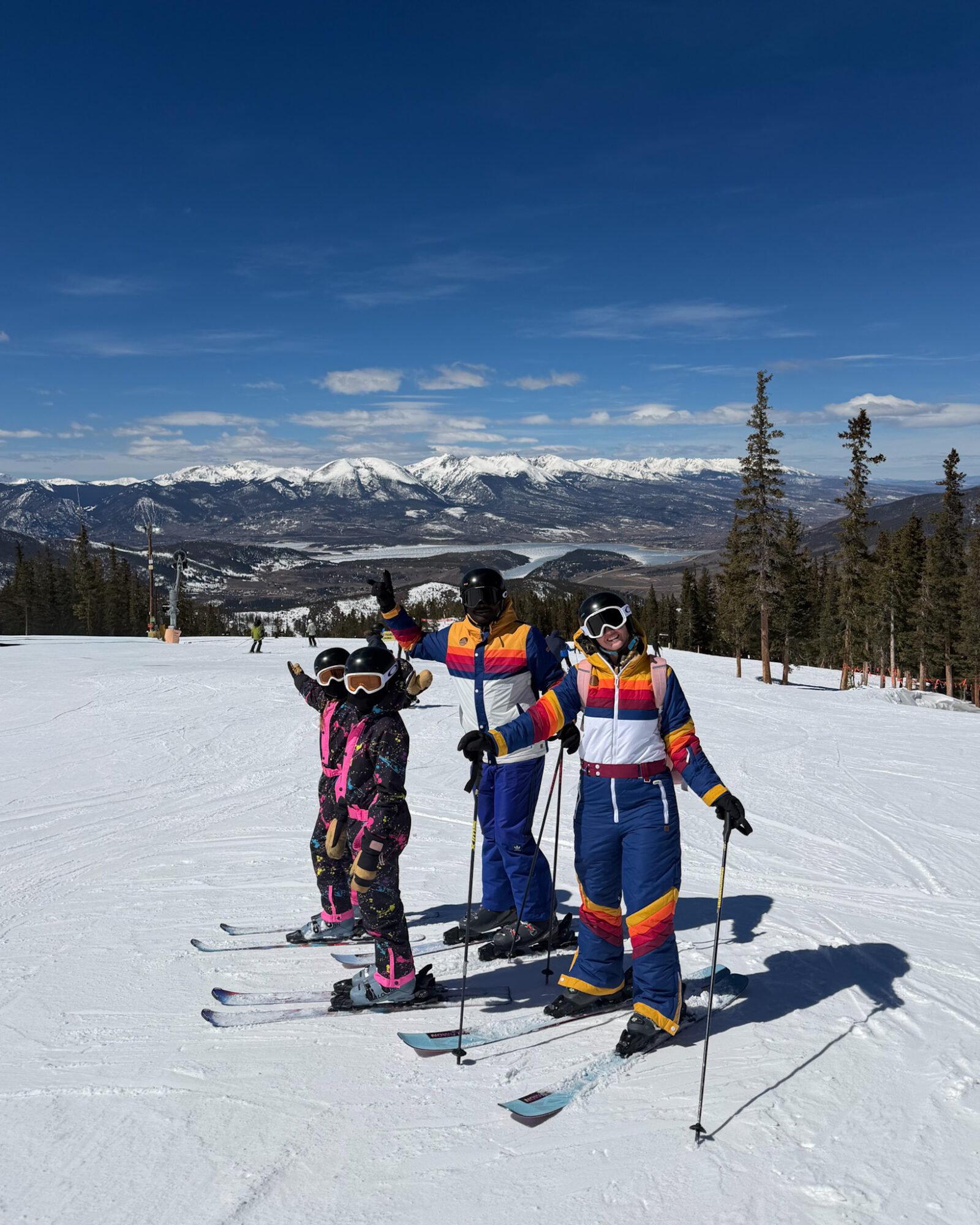 Group of children in colorful winter clothing skiing on snow with mountains and trees in background.