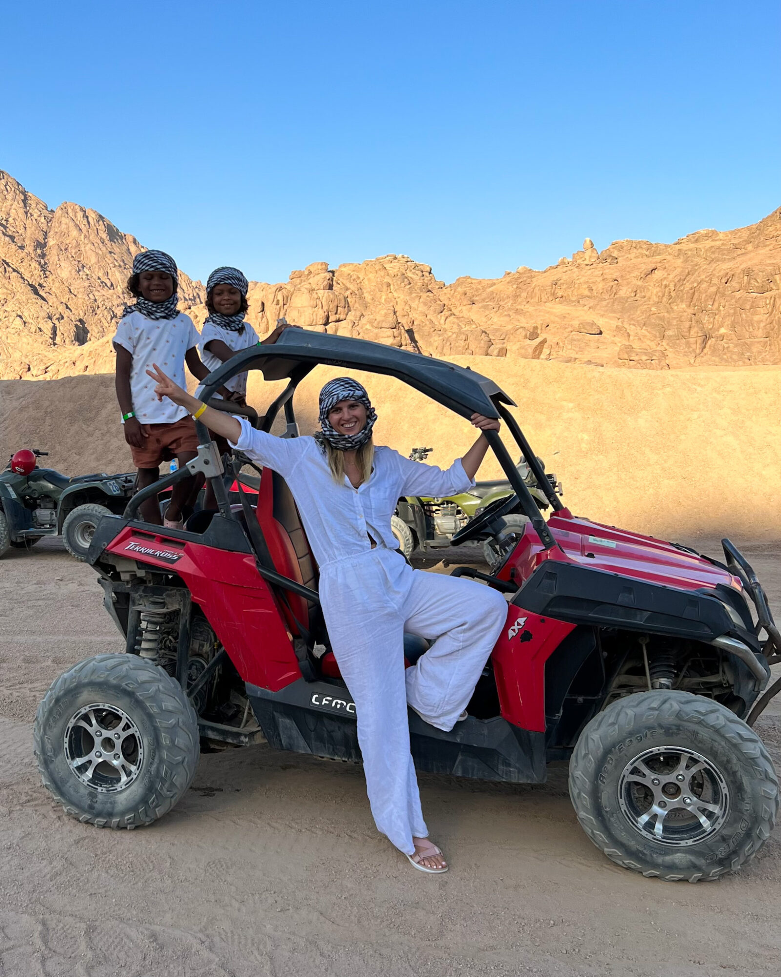 Three people in desert landscape with rocky hills, one sitting in a red off-road vehicle, two standing nearby, all wearing helmets.