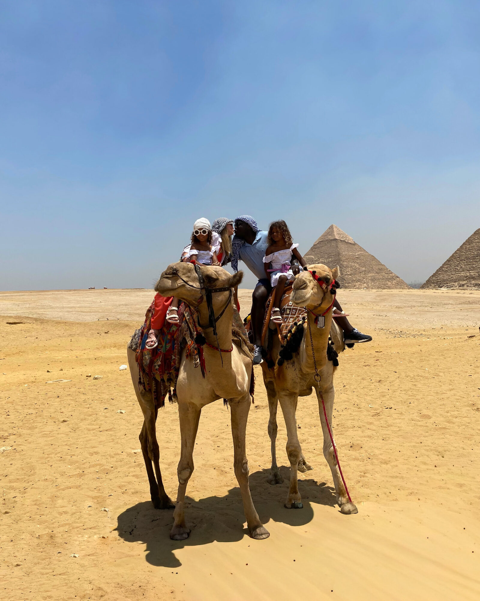 Three people riding camels in a desert with pyramids in the background under a clear blue sky.