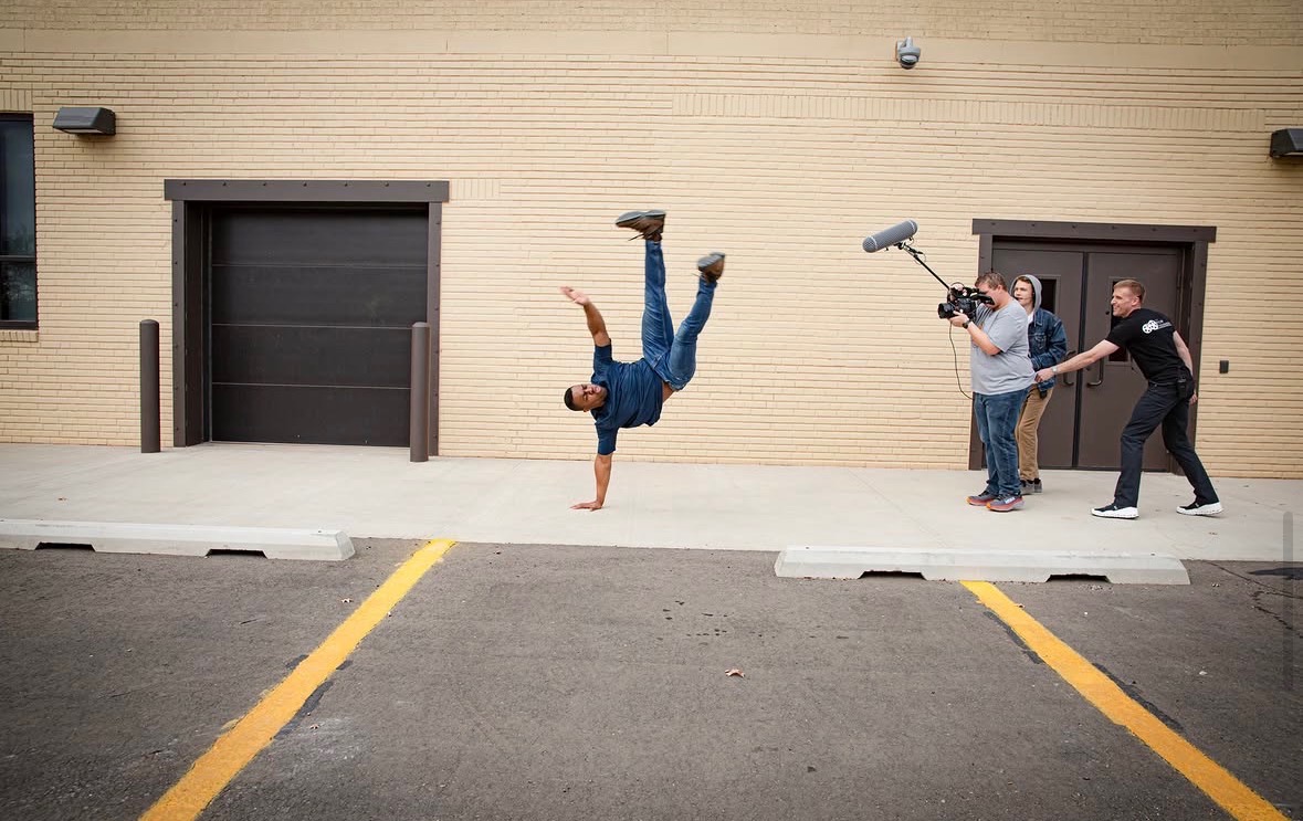 Person performing a handstand on the street with two camera operators filming nearby, against a beige building.