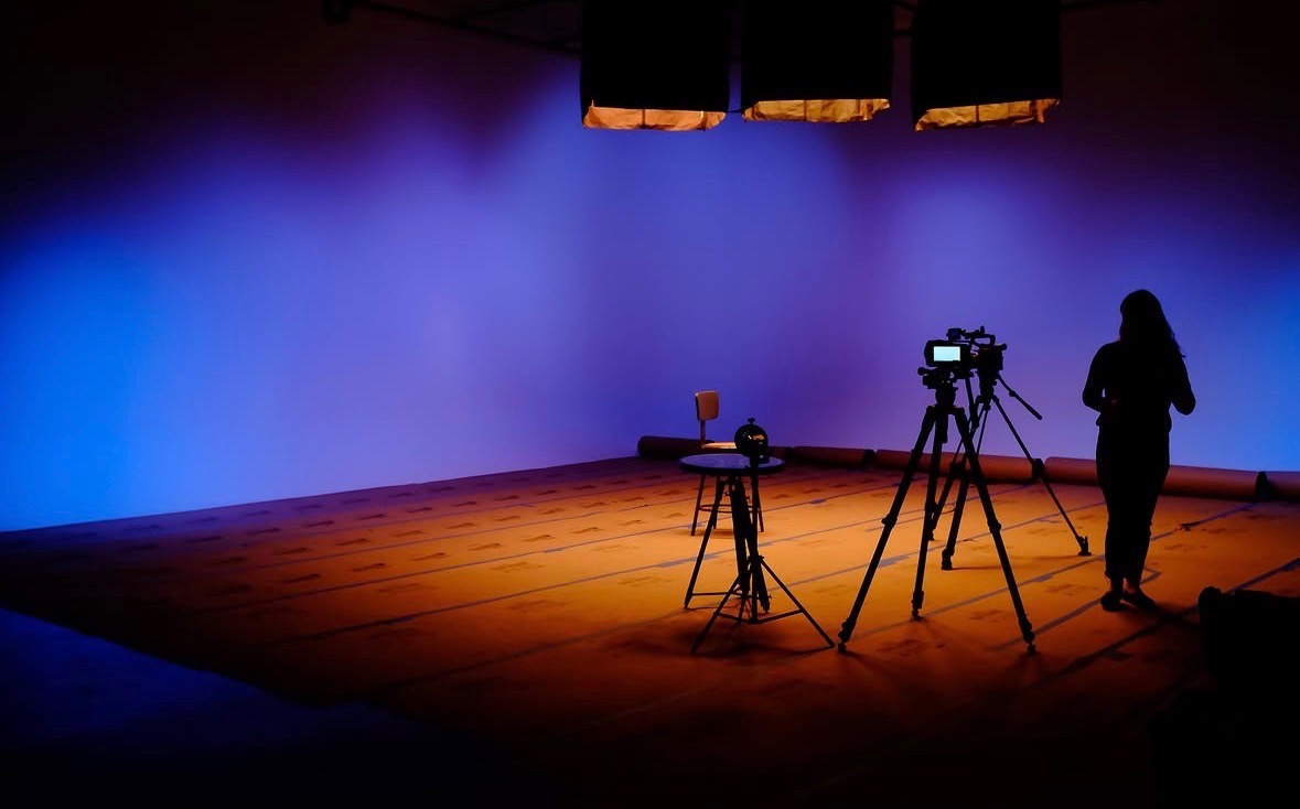 Stage with blue lighting, camera equipment, and a person preparing to film or photograph, with a wooden floor and curtains above.