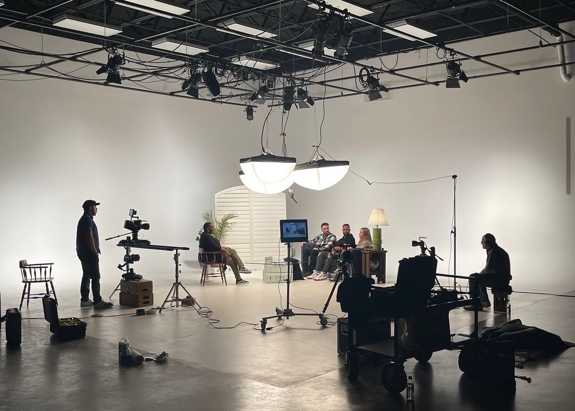 People in a studio with cameras, lights, and a monitor, filming a group seated on chairs, with a person operating equipment.