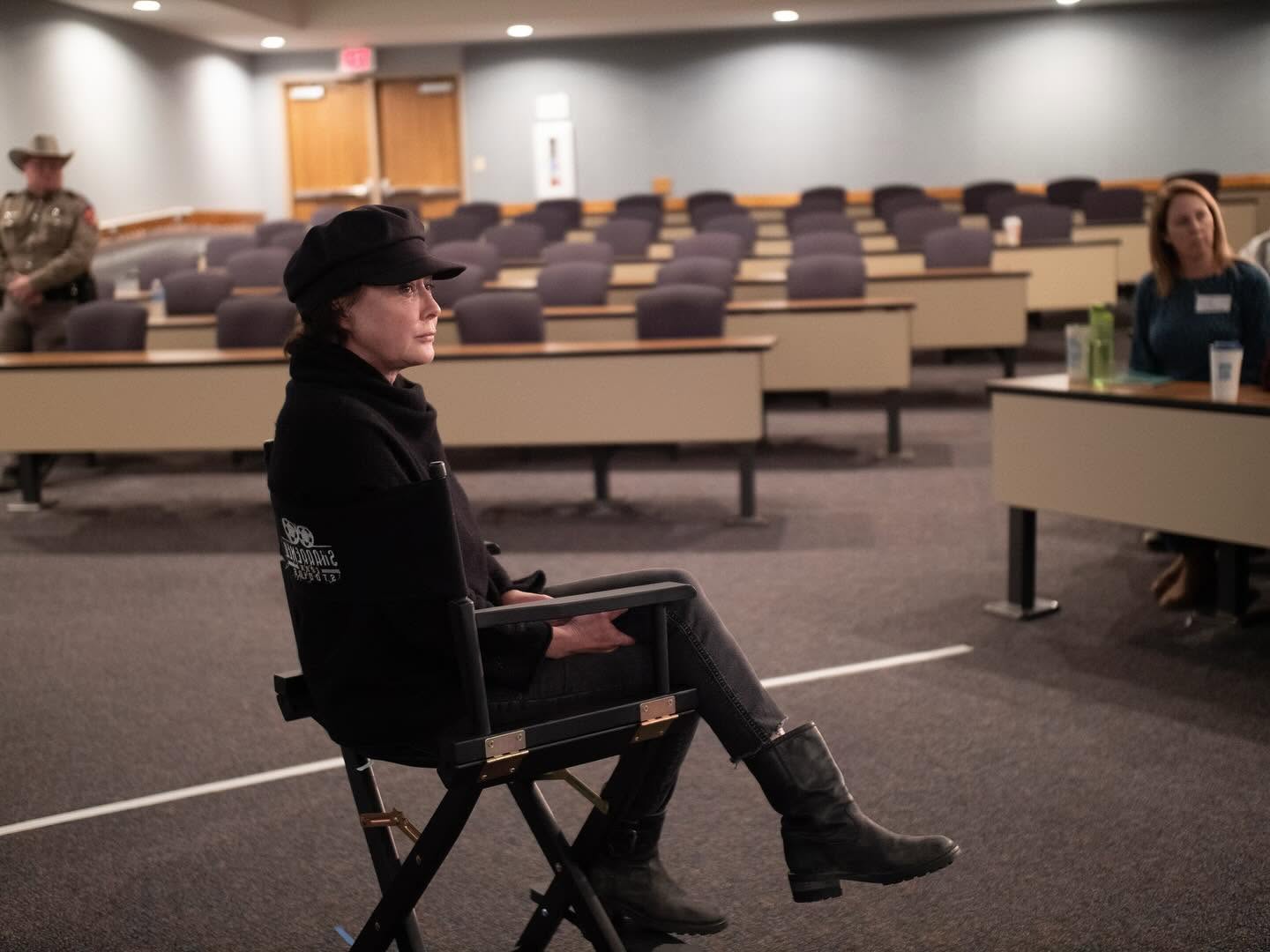 Woman sitting on a chair in an empty conference room with rows of seats and a few people present.