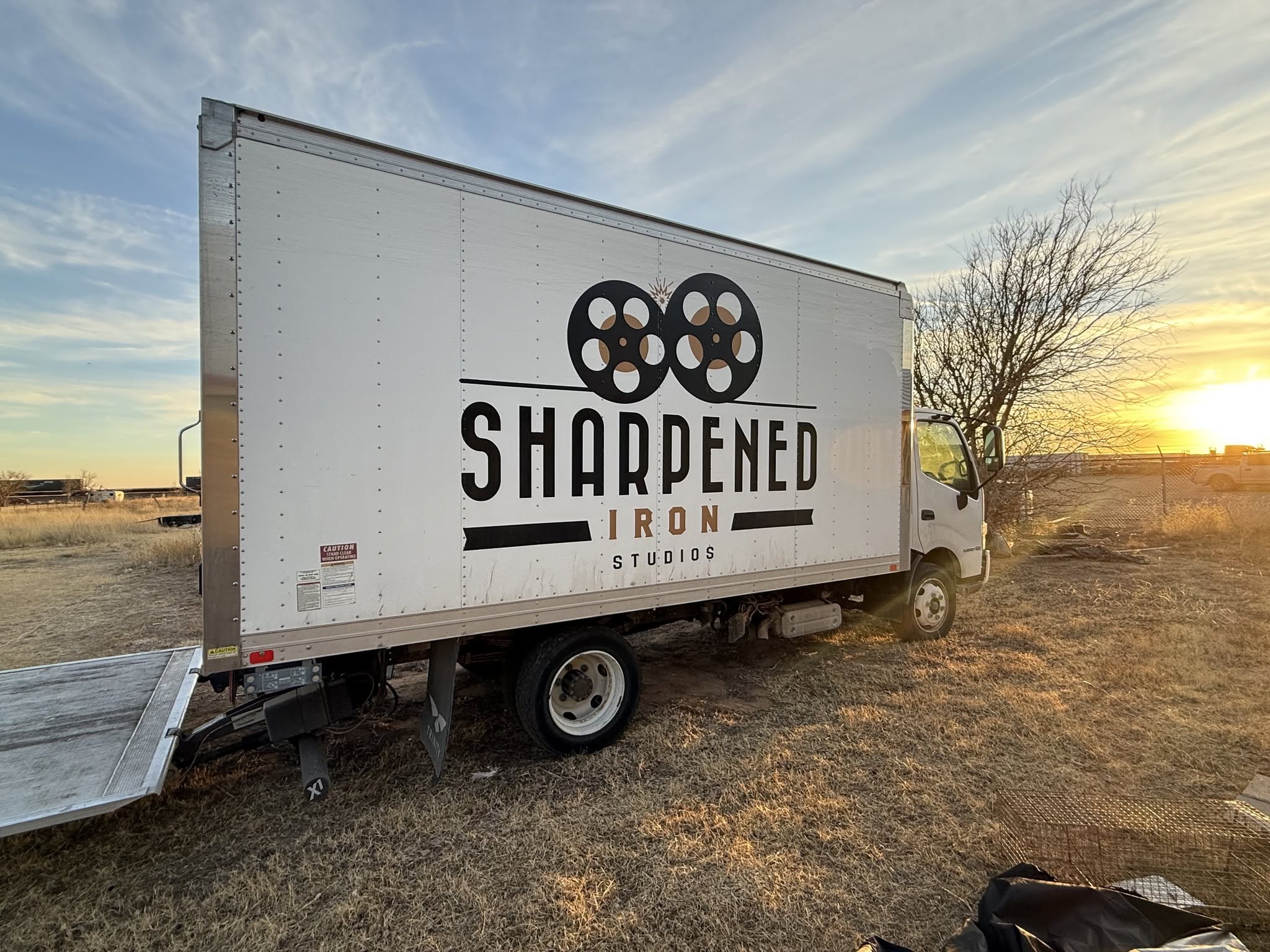 White truck with logo and text on side, parked on grassy field during sunset, with a tree and open sky in background.