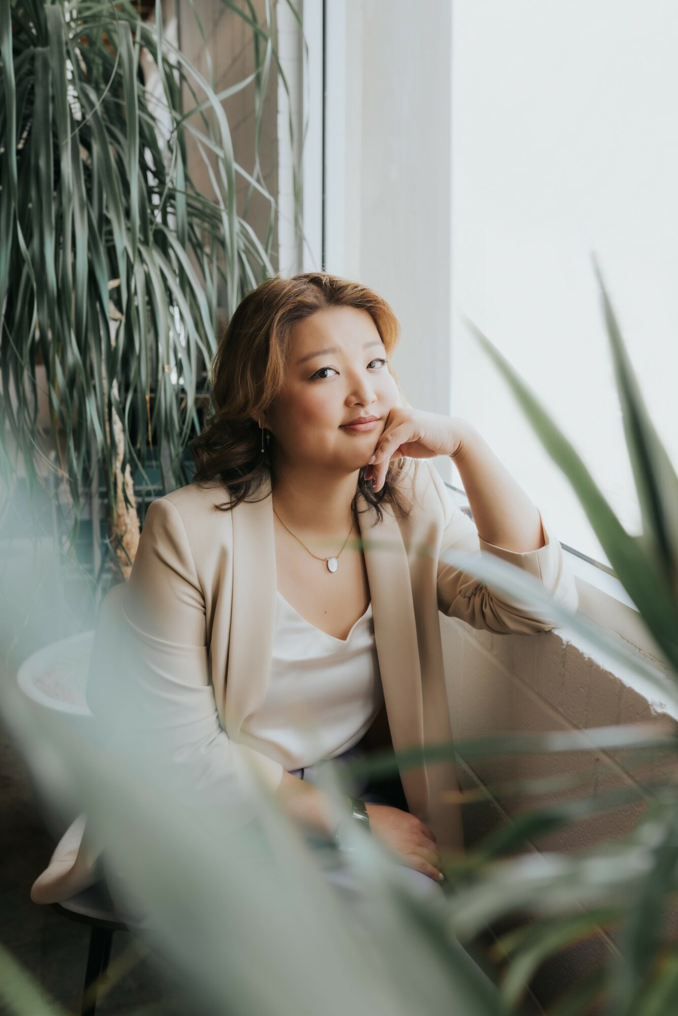 Woman with shoulder-length hair sitting by a window surrounded by plants, looking at the camera with a slight smile.