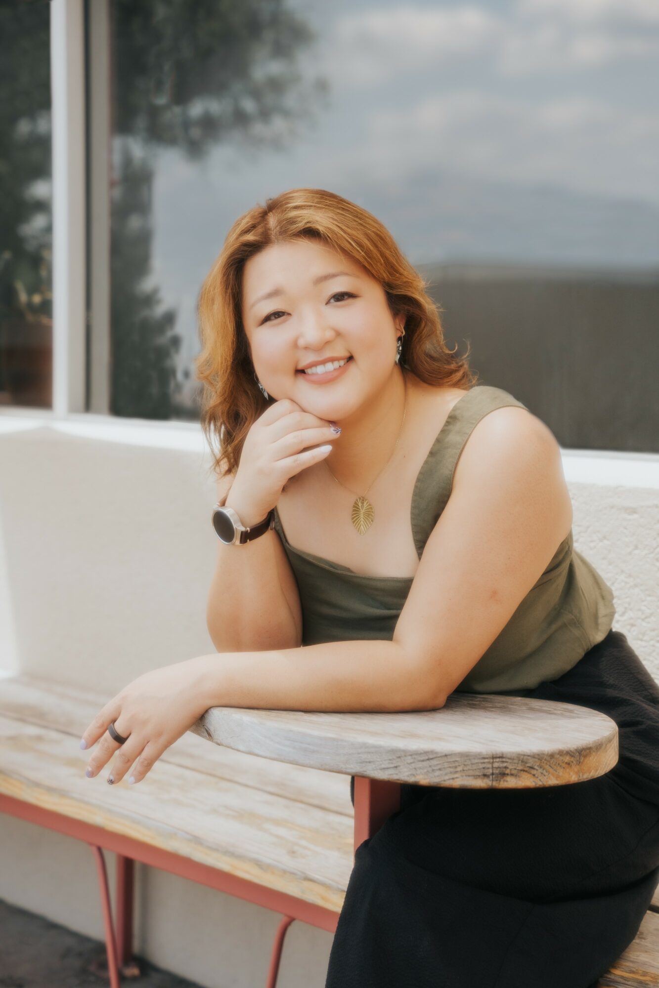 Woman with shoulder-length hair smiling, resting her chin on her hand, sitting at a table indoors with a window behind her.