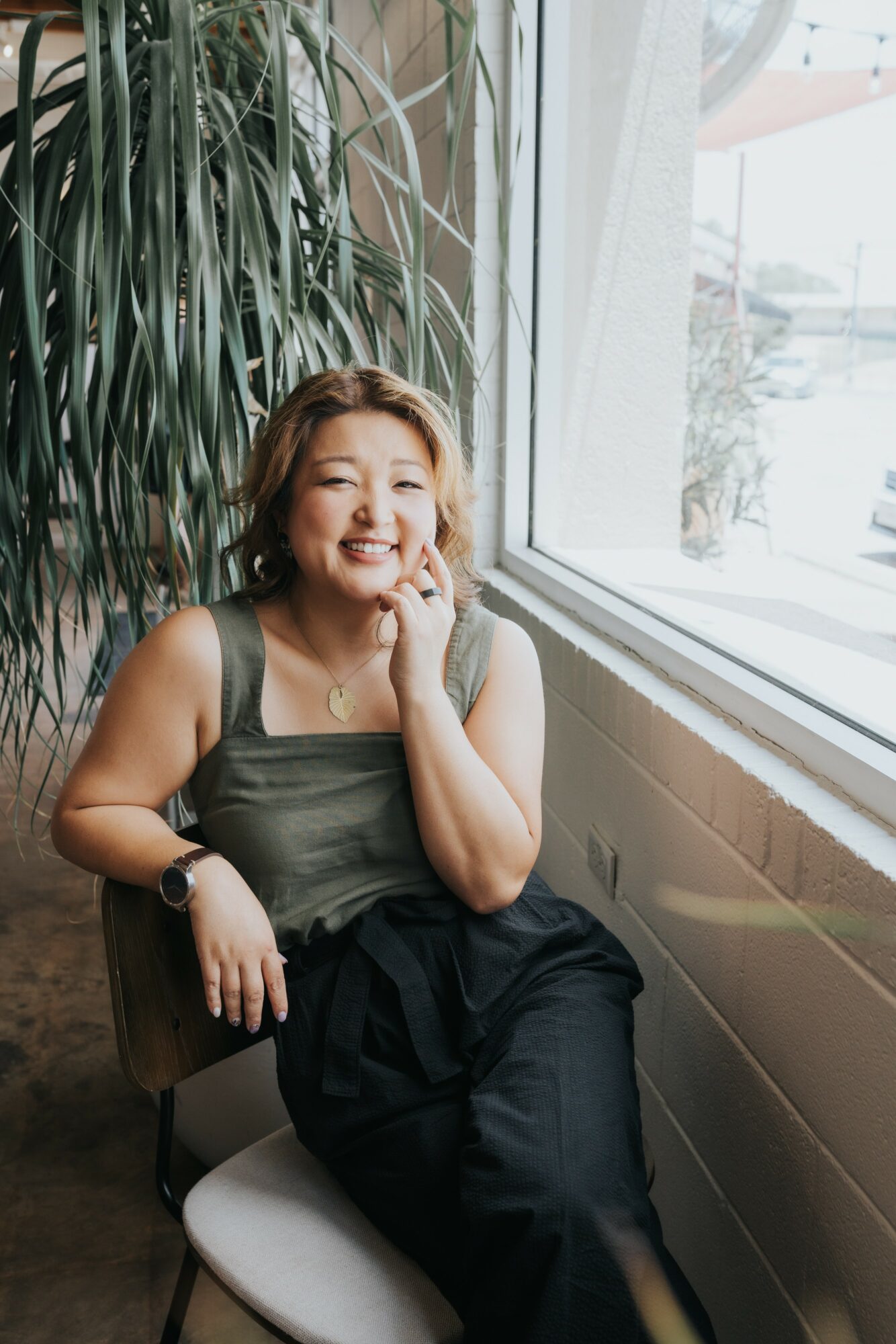 Woman sitting by window, smiling, hand on face, indoor setting with large plant, natural light, casual attire.