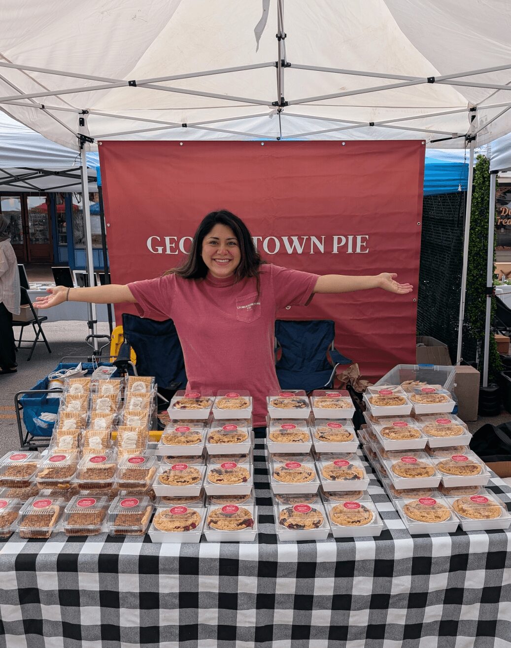 Woman standing behind a table with pies at an outdoor market stall, arms outstretched, smiling, with a red sign in the background.