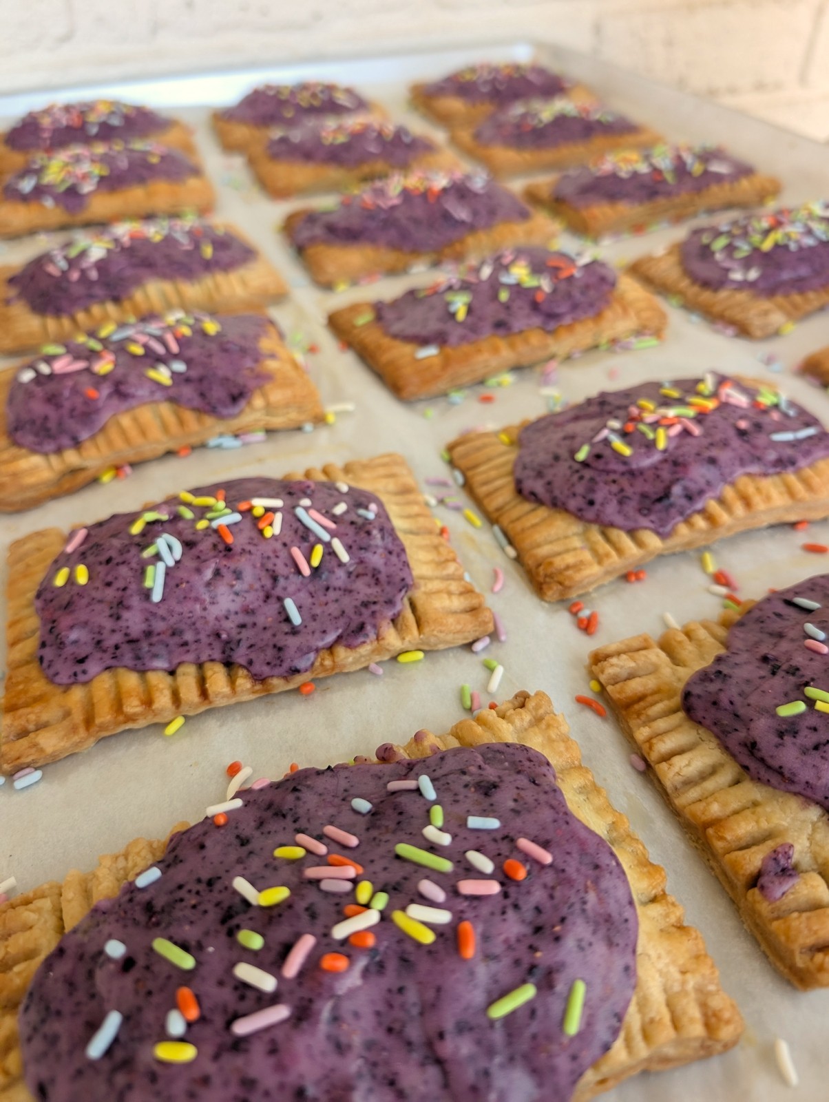 Rectangular cookies with purple frosting and colorful sprinkles on a baking sheet, some with chopped nuts, arranged in rows.