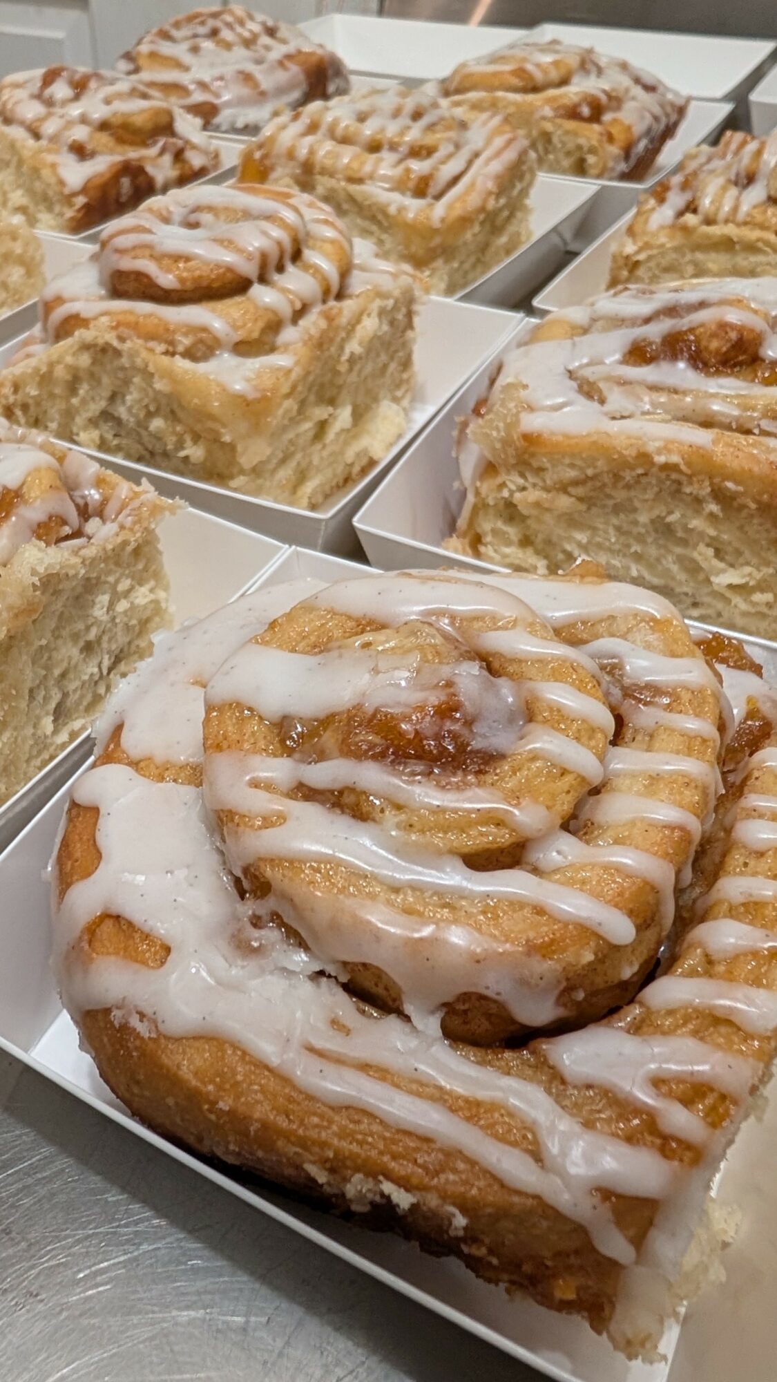Several cinnamon rolls with icing in white boxes, some cut open showing filling, on a gray surface.