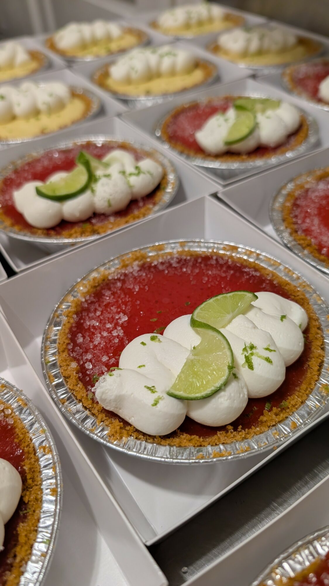 Multiple fruit tarts with lime slices, whipped cream, and red fruit glaze in individual containers.