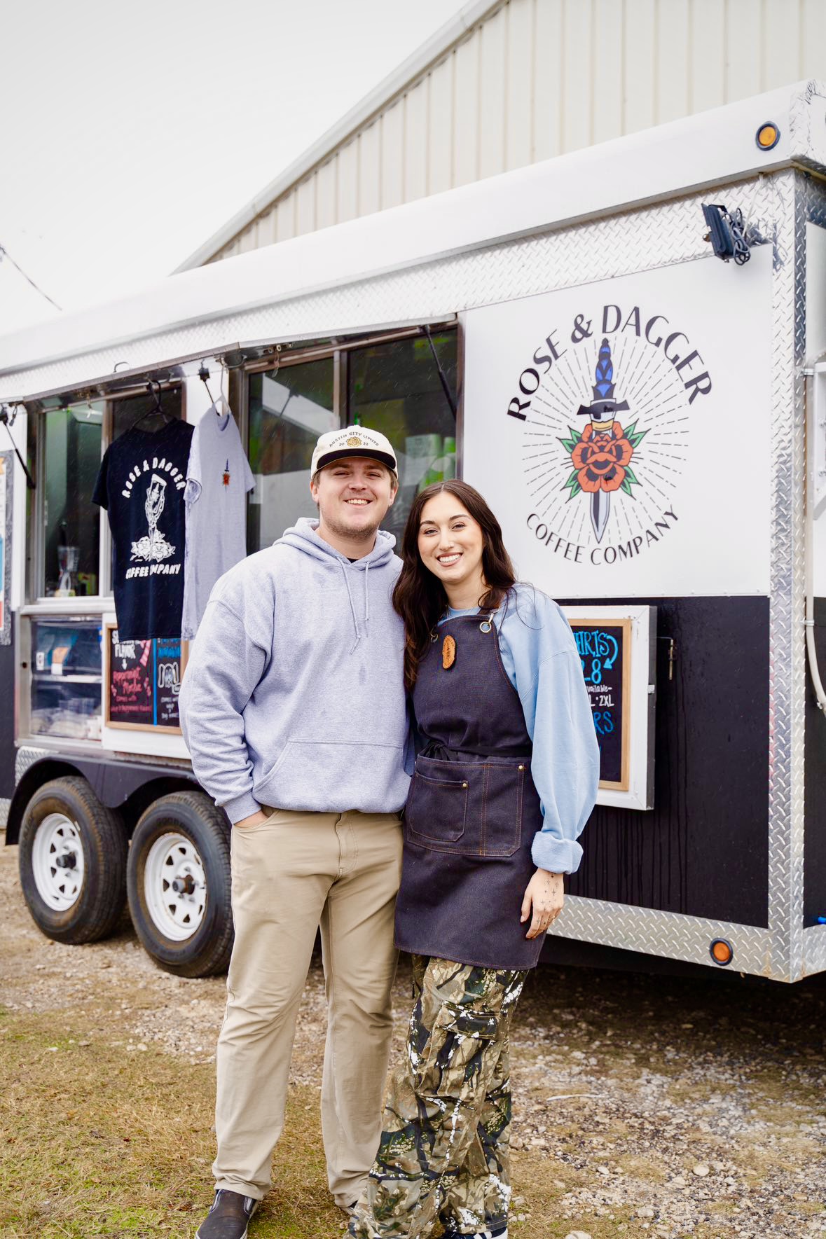 Two people stand in front of a food truck with a logo and text, smiling at the camera.