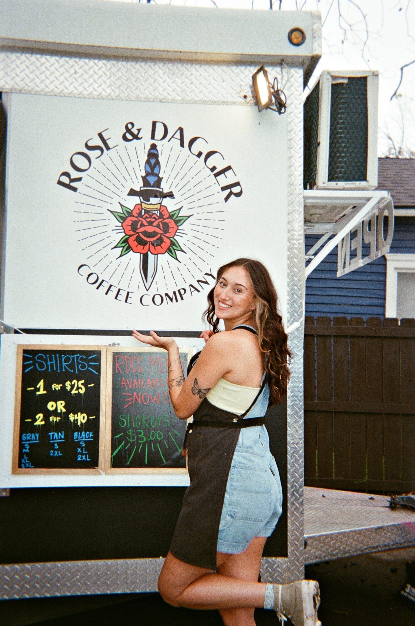 Young woman with long hair kneeling in front of a coffee truck, smiling, with a logo and menu board visible.