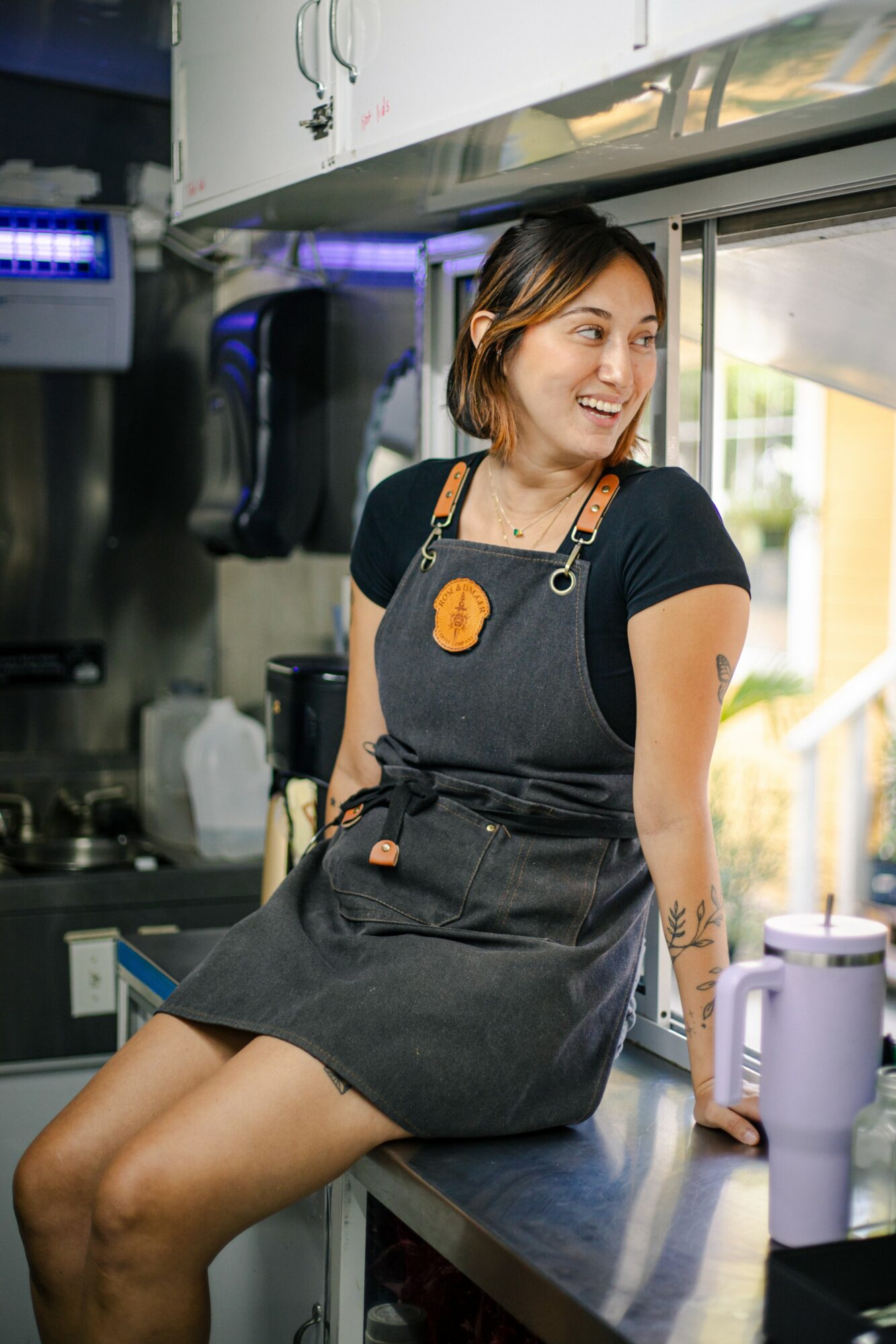 Woman in apron sitting on kitchen counter, smiling, with kitchen appliances and window behind her.