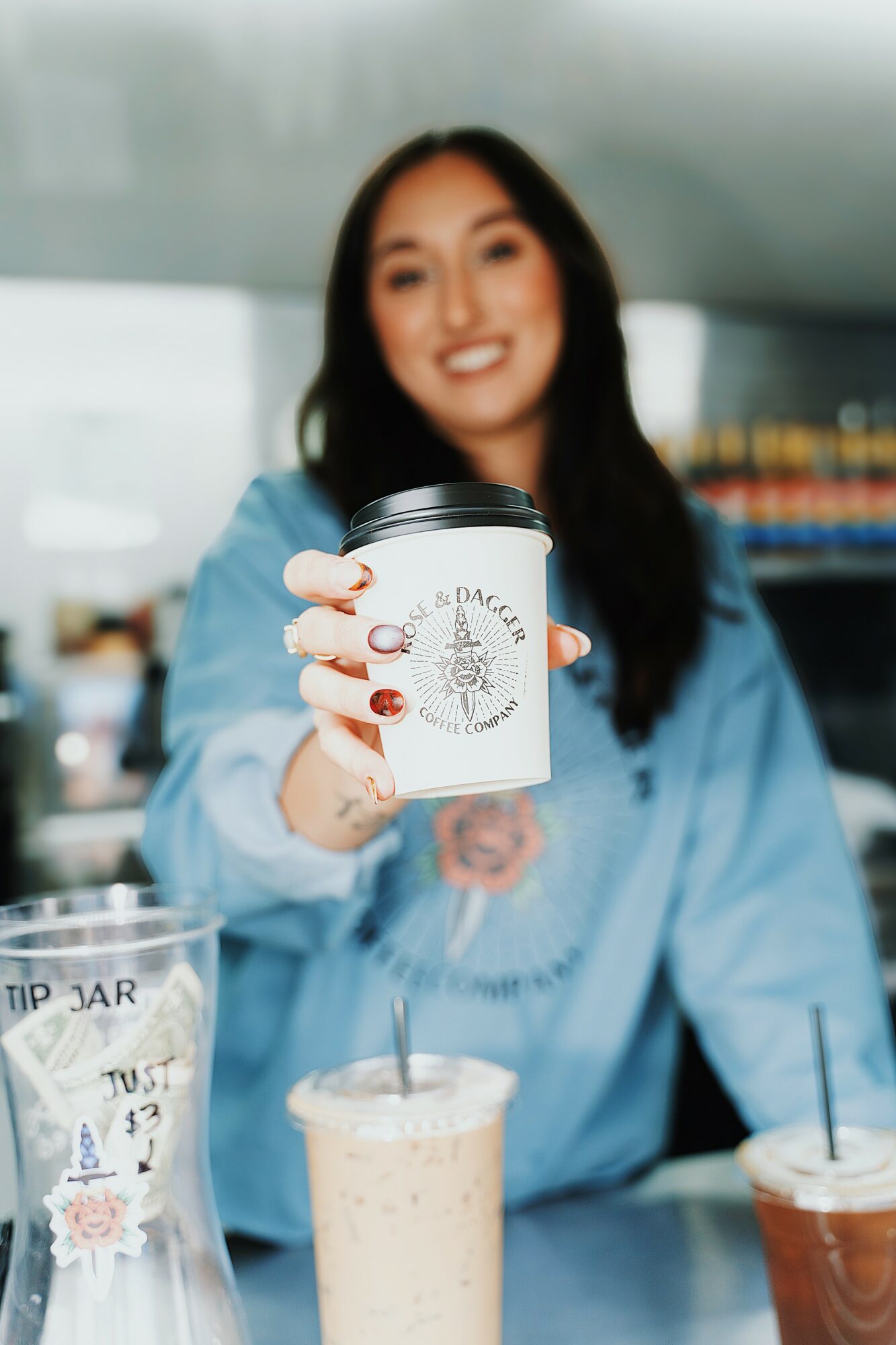 Woman smiling and holding a coffee cup in a cafe, with a tip jar and drink on counter, blurred background.
