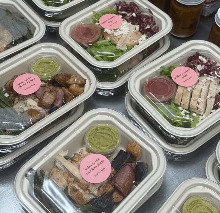 Multiple clear plastic food containers with salads and sides, some with pink labels, on a table surface.